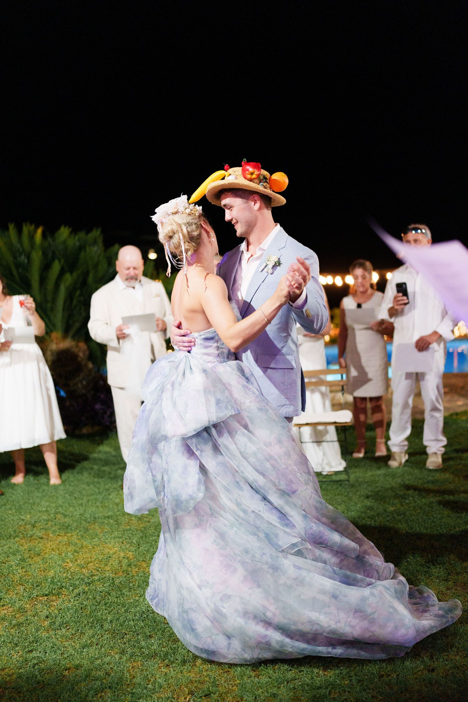 Couple shares first dance outdoors at night, bride in floral gown, groom in gray suit with fruit hat, guests watching.
