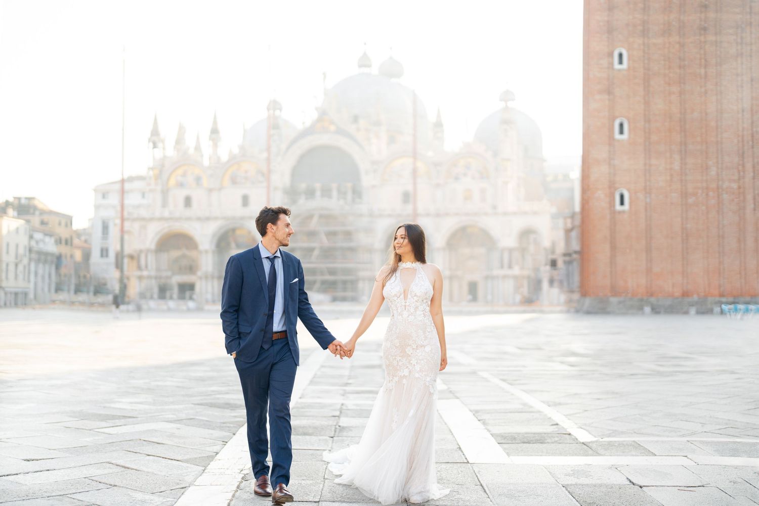 A couple in wedding attire walks hand in hand across a grand European plaza with historic architecture in the background.