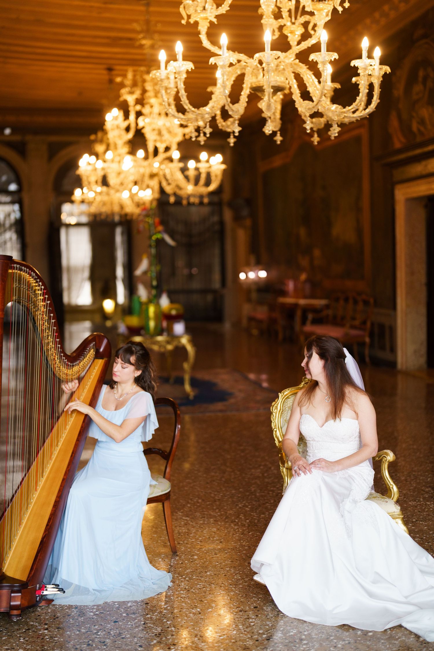 Two musicians in elegant dresses perform on harps in an ornate ballroom with crystal chandeliers.