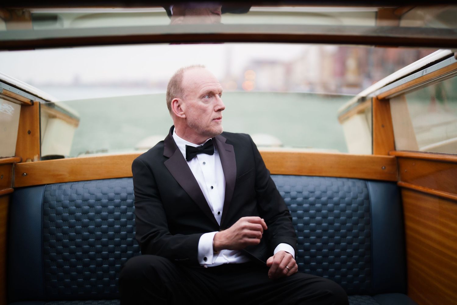 A person in formal attire sits on a dark quilted leather bench seat in what appears to be a luxury vehicle interior.