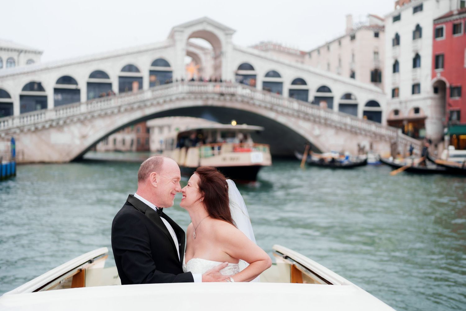 A romantic couple shares an intimate moment while riding in a gondola along Venice's iconic canals with bridges in view.