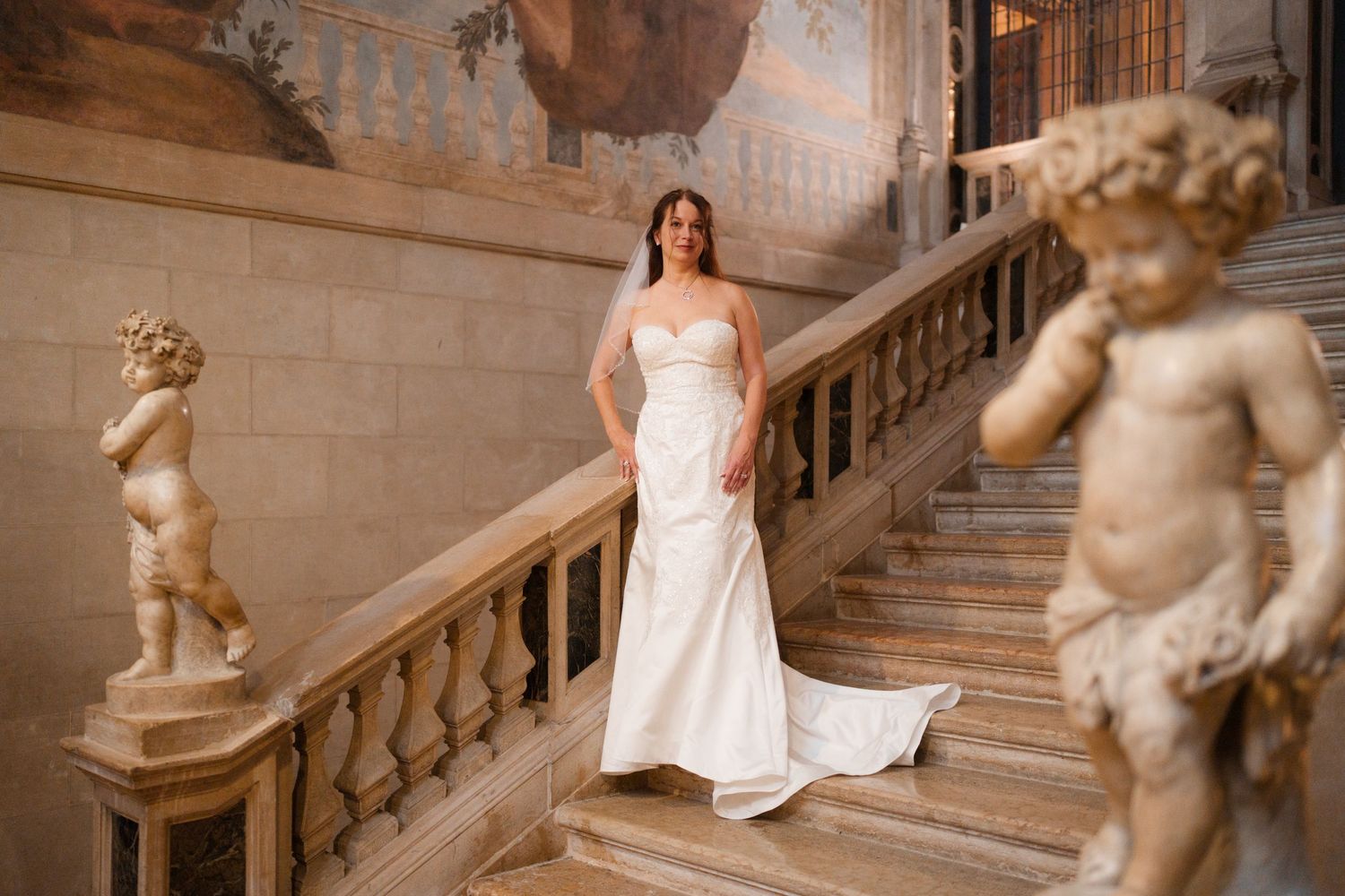 A bride in a white wedding dress poses elegantly on an ornate marble staircase surrounded by classical statues.