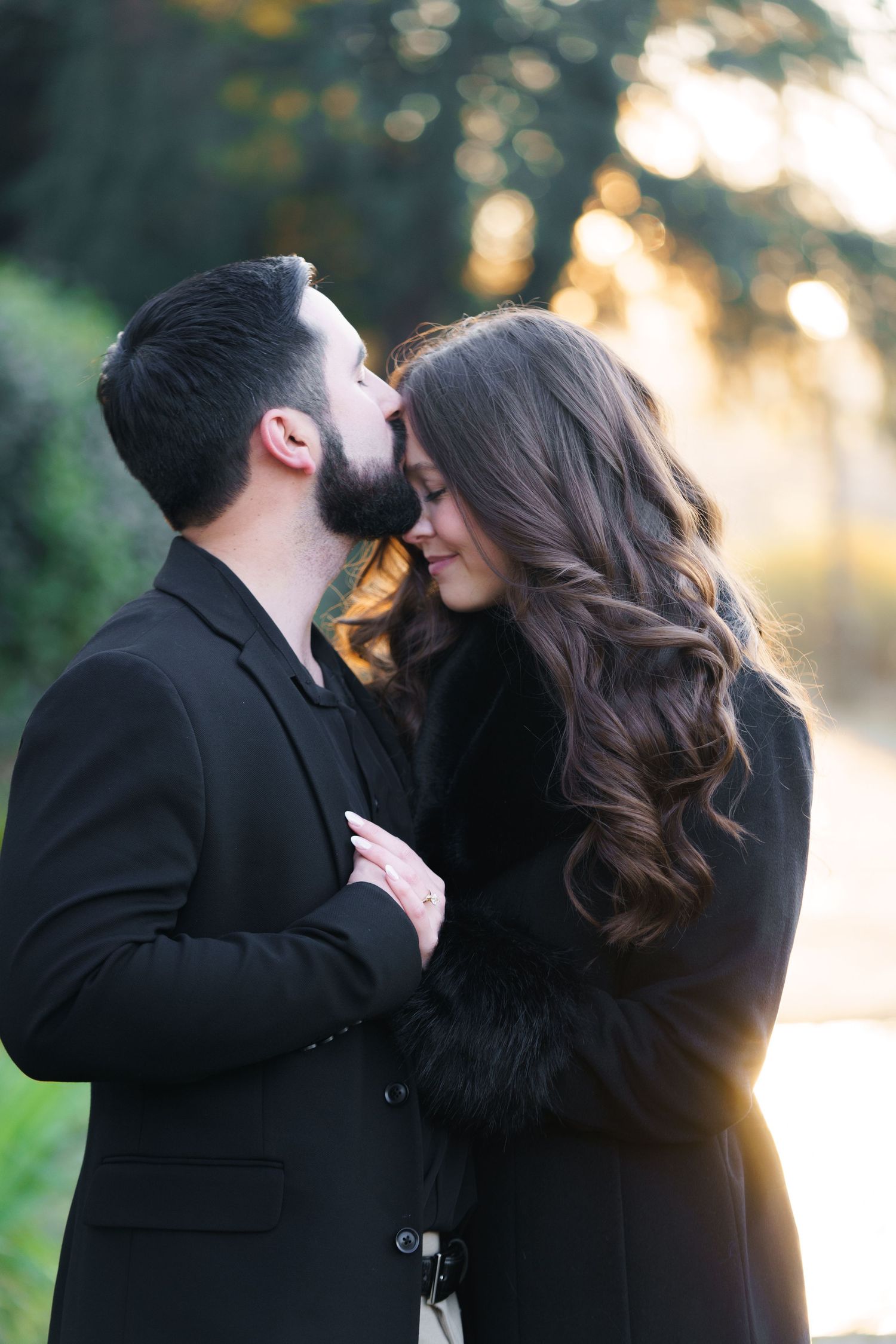 A romantic couple in black coats share intimate moments at sunset in a natural outdoor setting.