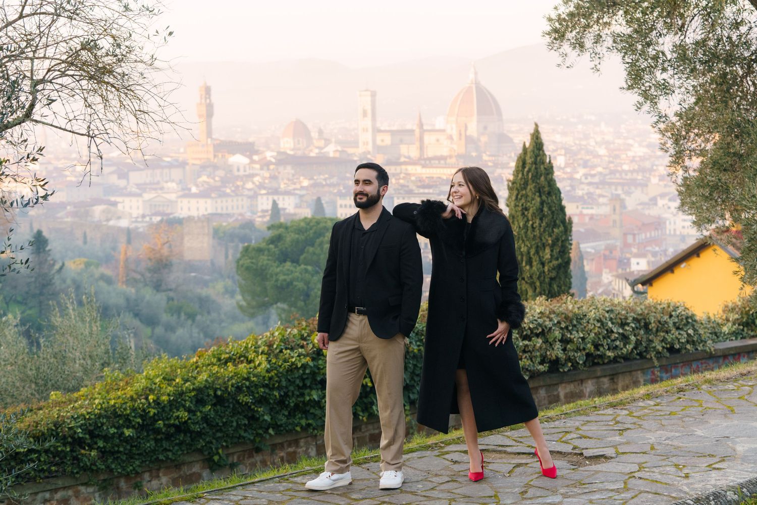 Two people stand on a scenic overlook with Florence's iconic skyline and Duomo cathedral visible in the misty background.