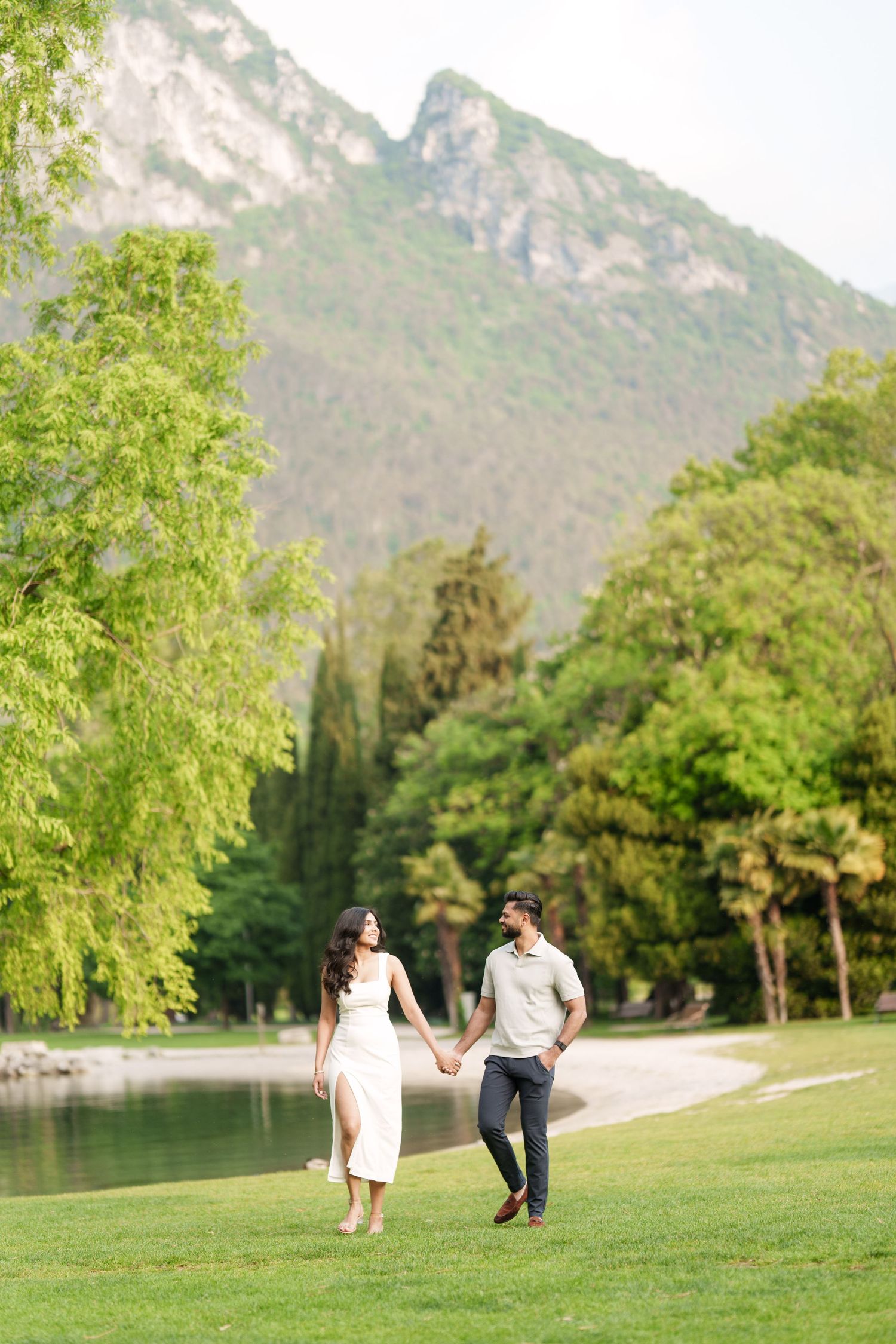 Couple walking hand in hand on green lawn near mountain lake with scenic peaks and lush trees in background.