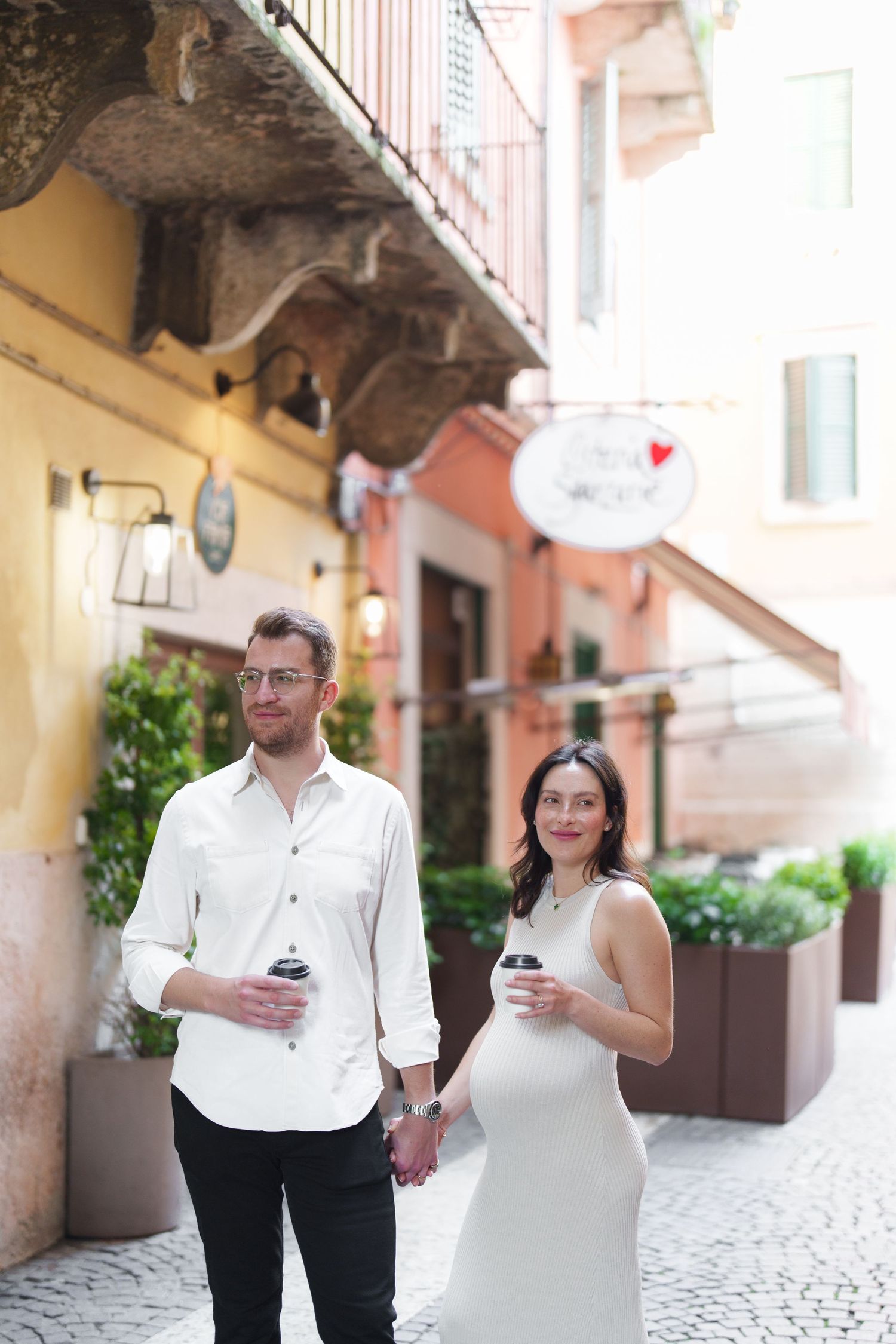 A couple in white attire walking through a charming European alleyway while looking at their phones.
