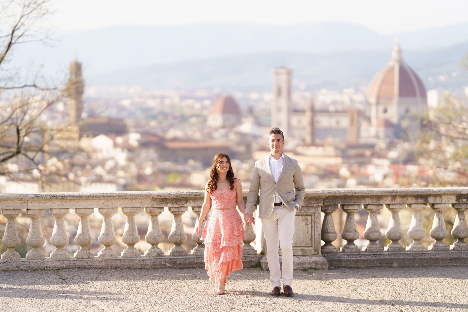 A couple in elegant attire poses on a balcony overlooking the historic Florence skyline with the Duomo cathedral visible.