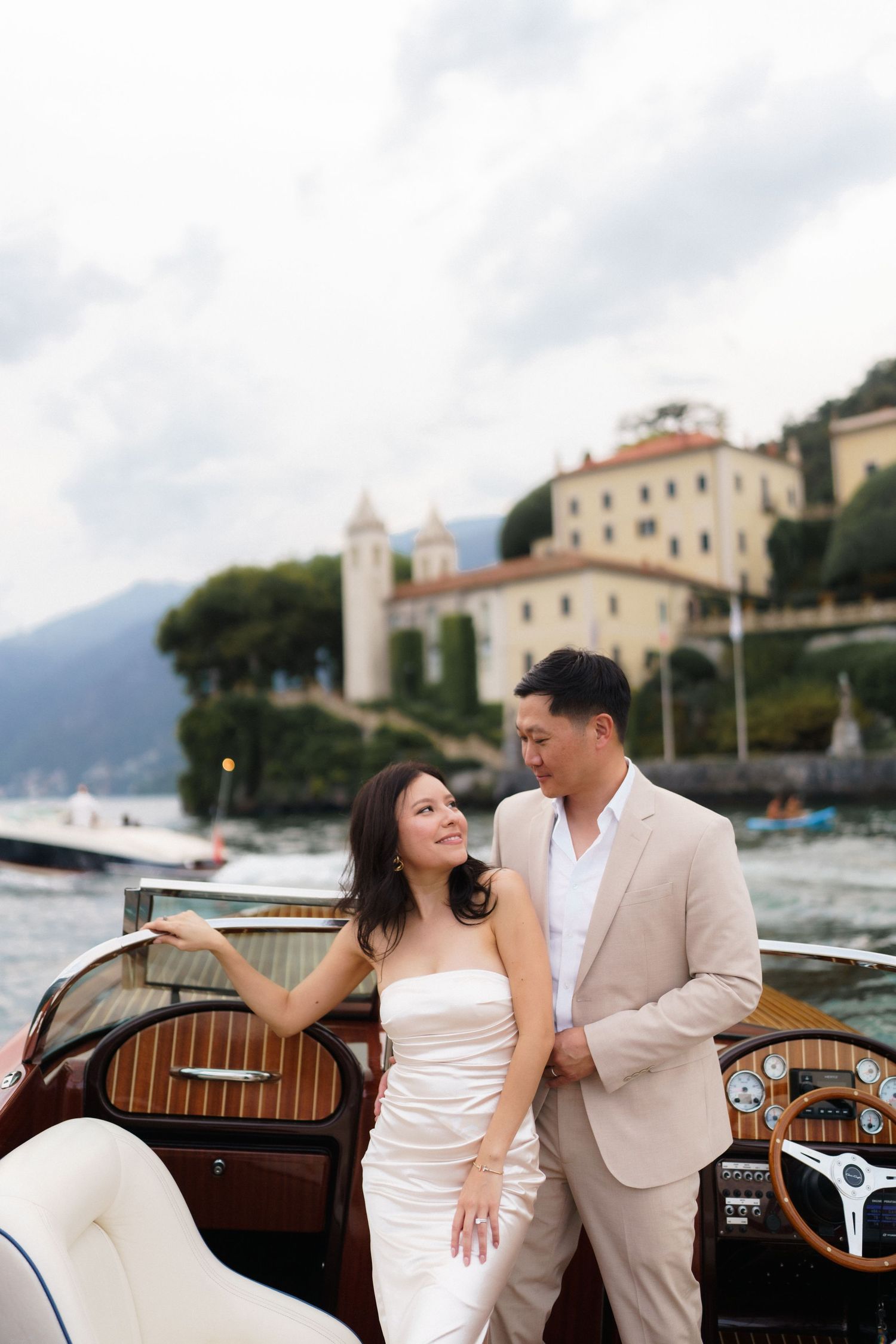 A romantic couple shares intimate moments on a classic wooden boat along Lake Como's shoreline with Italian villas in view.