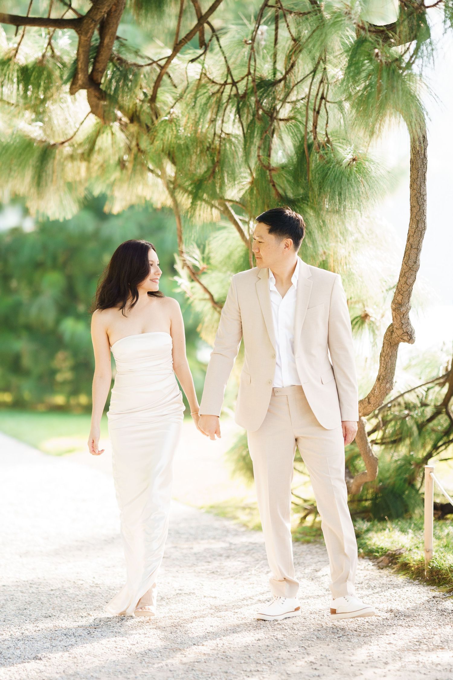A couple in white outfits walks together beneath pine trees on a sunlit path in a romantic outdoor setting.