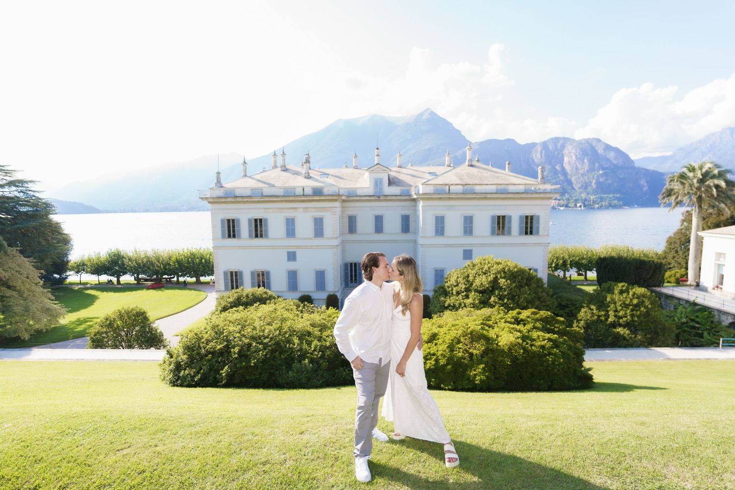 A couple in wedding attire walks along a scenic path with mountains and a white mansion in the background.