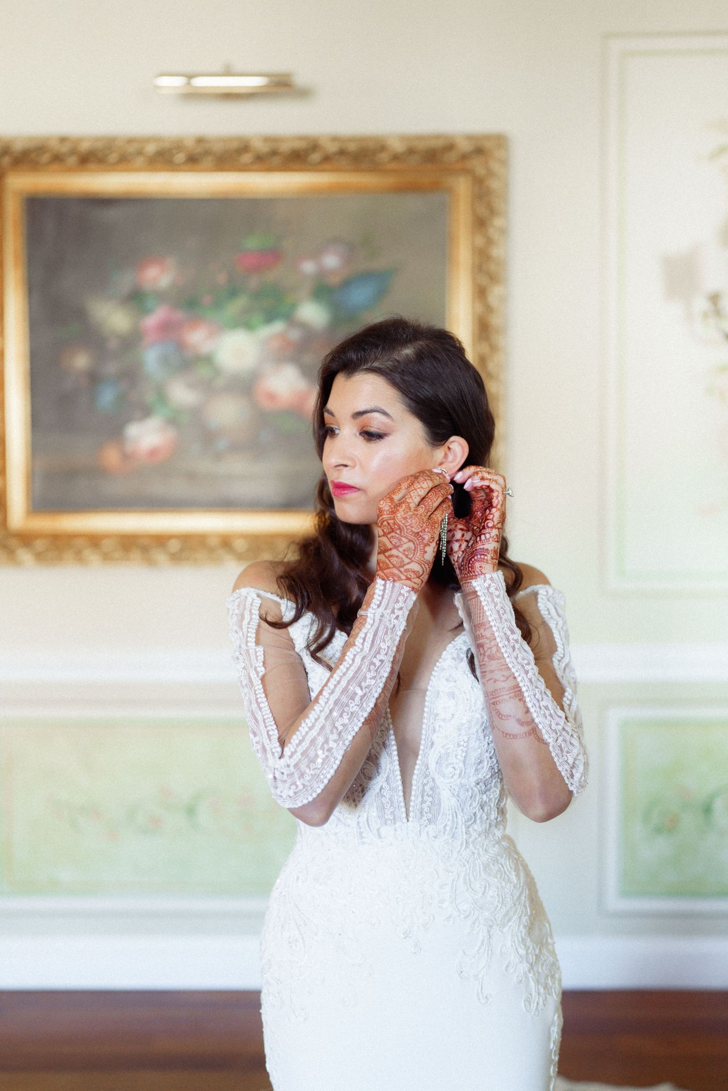 Bride in white lace dress showing traditional henna decorated hands in elegant indoor setting.