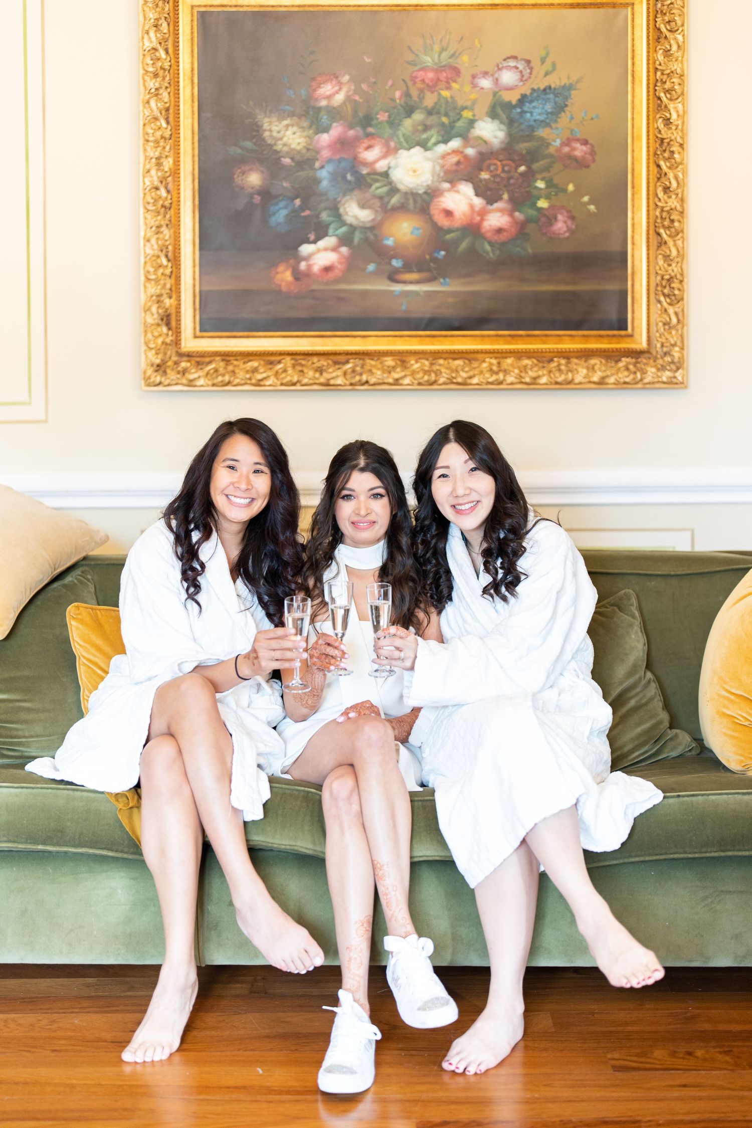 Group sitting on vintage green couch wearing white outfits in elegant room with gold-framed paintings.