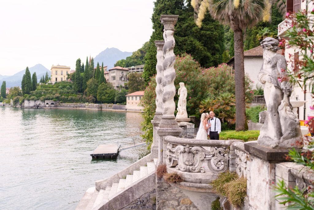 Stone statues line waterfront terrace of Italian villa with lake and mountain views.