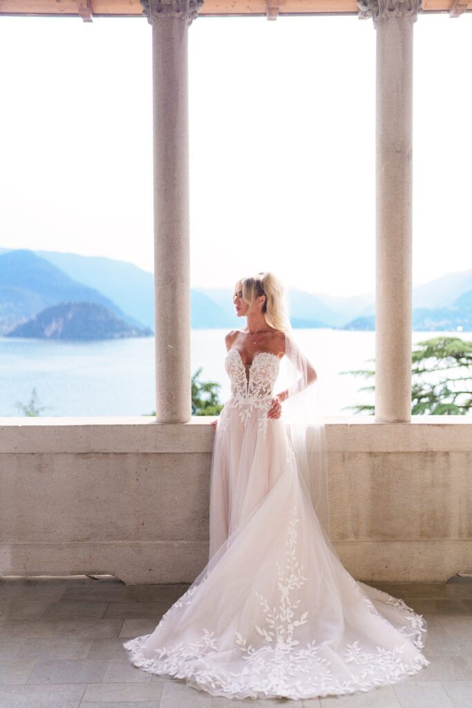Elegant white wedding dress with lace details and long train photographed against a columned terrace overlooking mountains.
