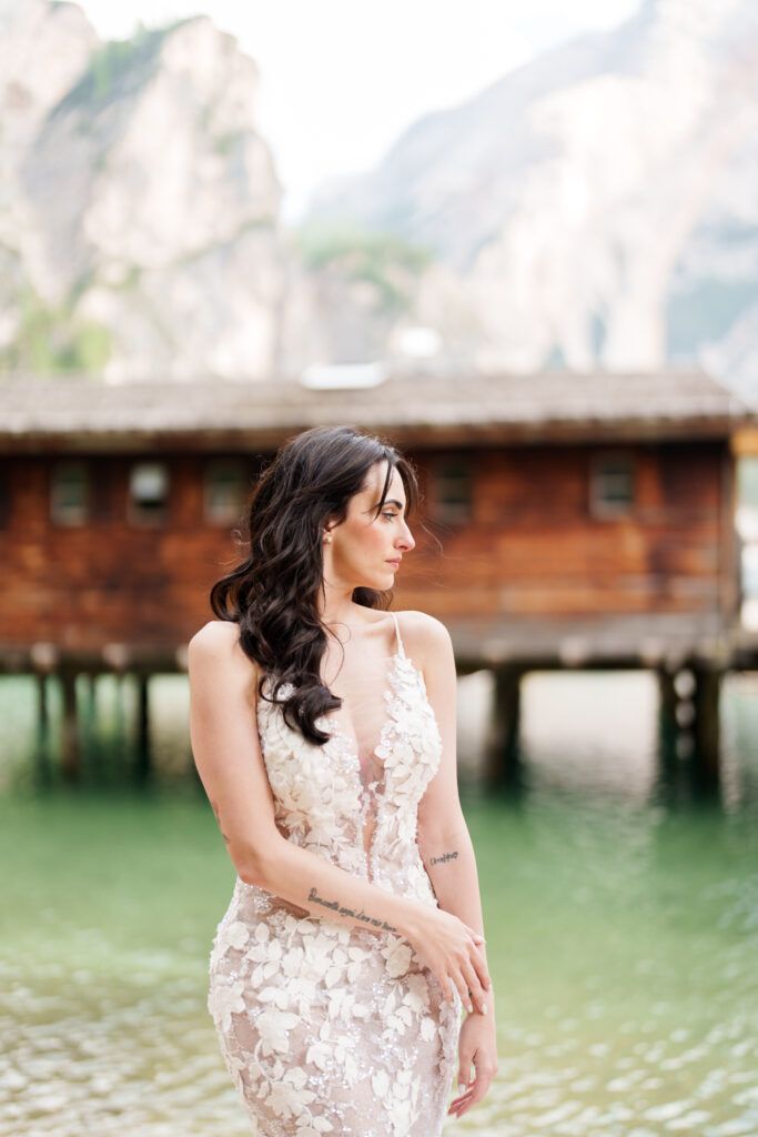 A bride in an elegant lace gown poses by a wooden dock on a serene mountain lake.