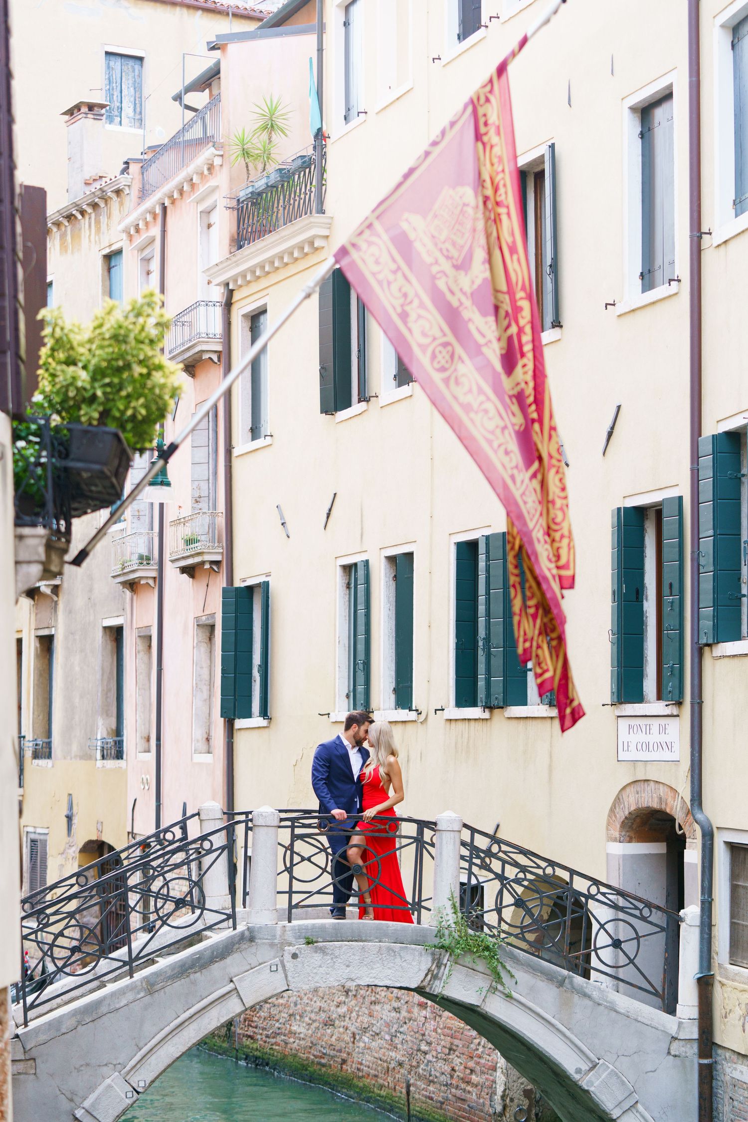 A narrow Venetian canal with ornate bridges and traditional buildings featuring green shutters and red flags.