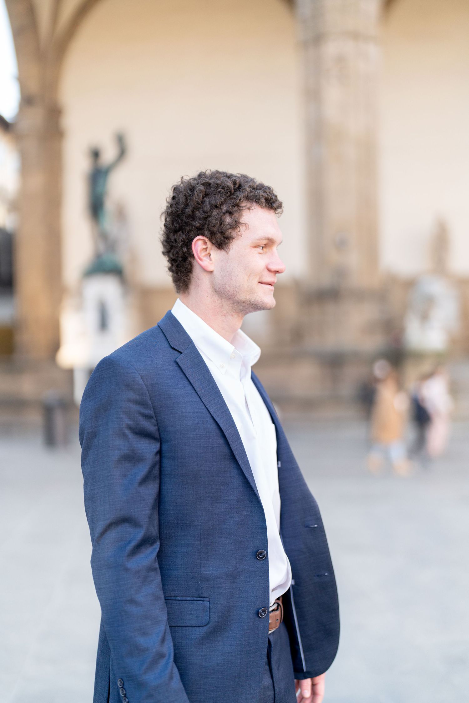 A person in a navy suit and white shirt stands in an outdoor plaza with architectural elements in the background.