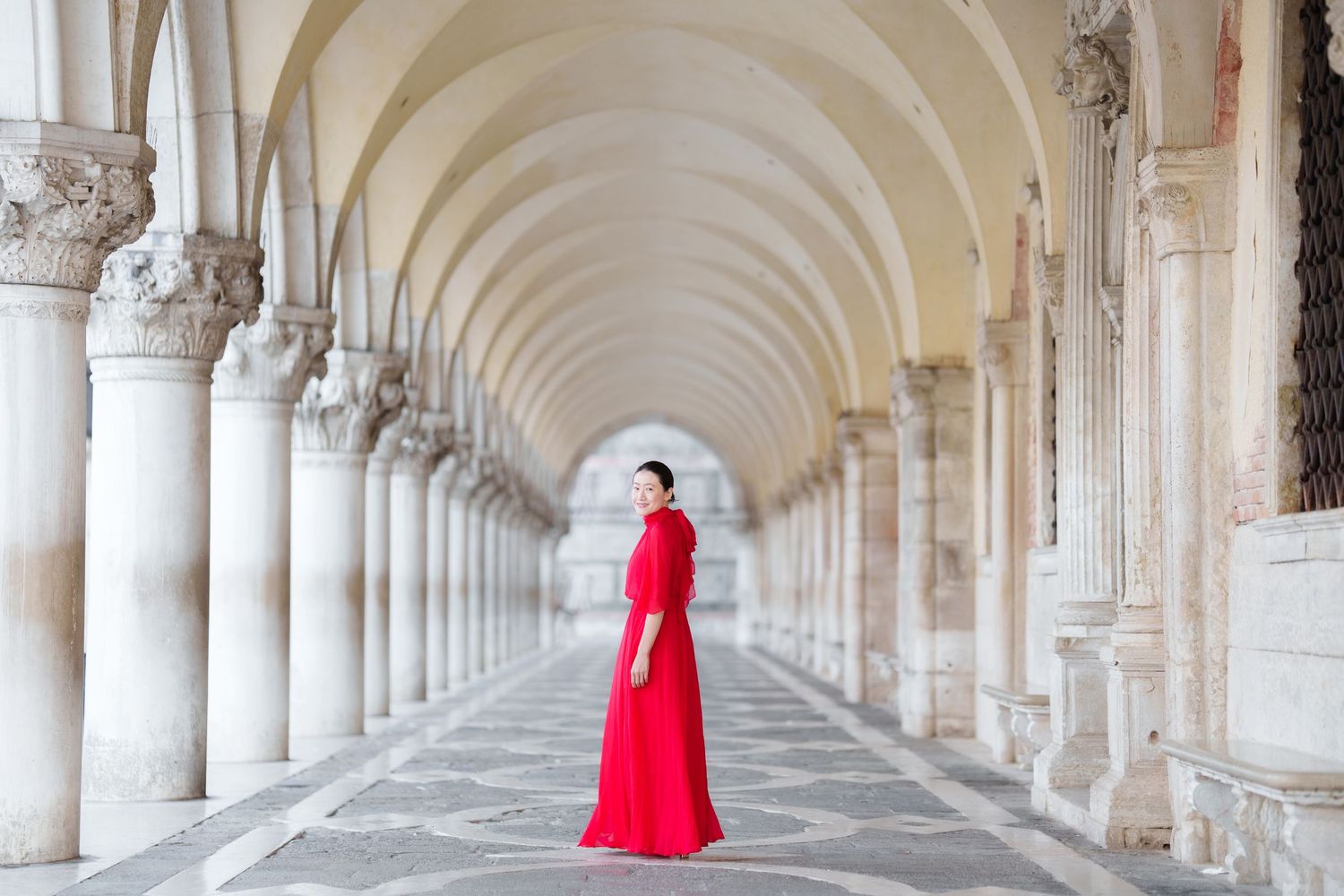 Woman in red gown standing in elegant marble colonnade arcade in Venice, Italy.