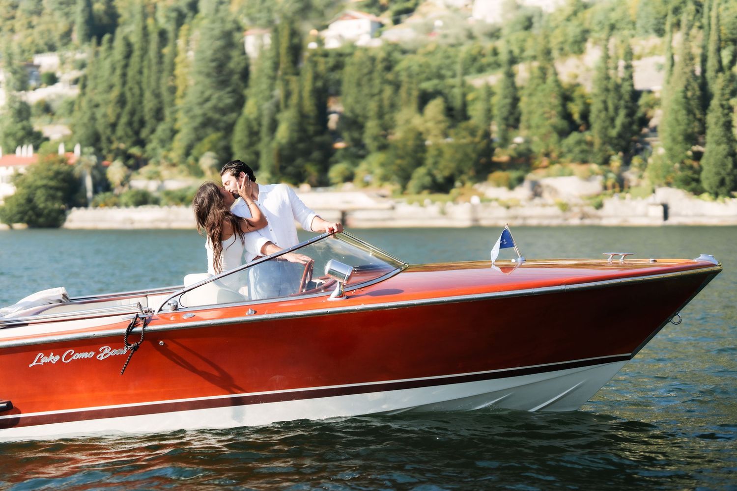 Couple kissing on a classic red wooden speedboat on Lake Como, surrounded by lush green trees.