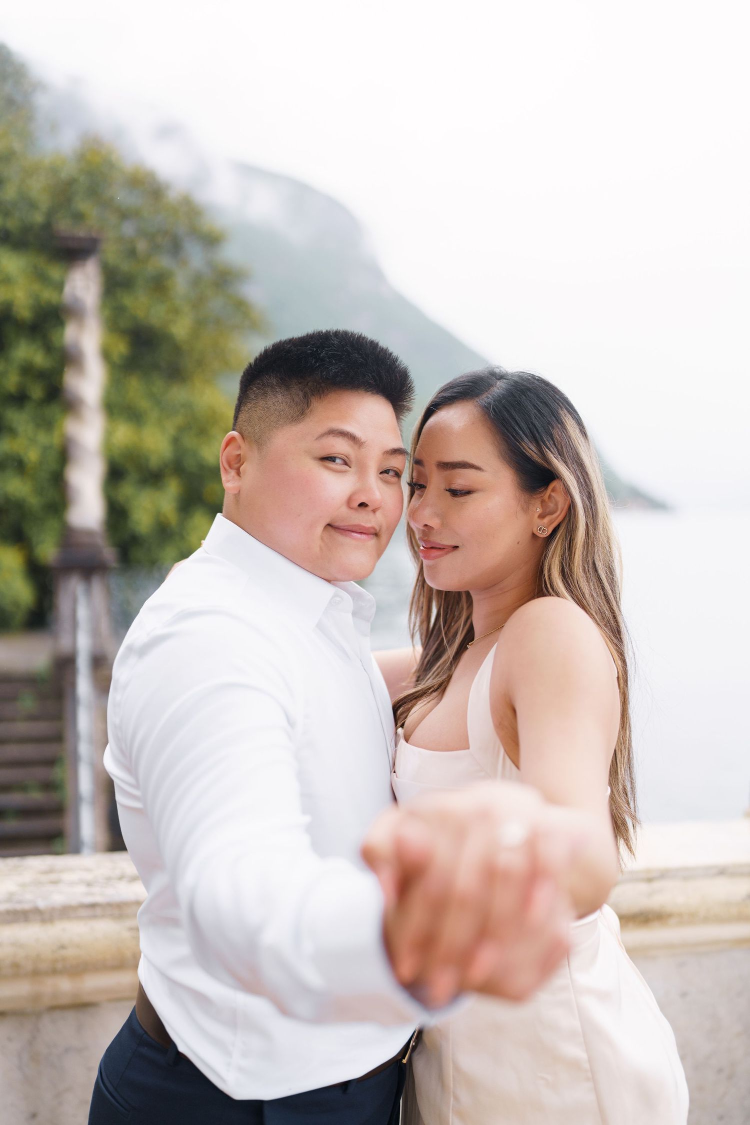 Couple sharing a romantic embrace outdoors near a scenic lakeside balcony with misty mountains in the background.