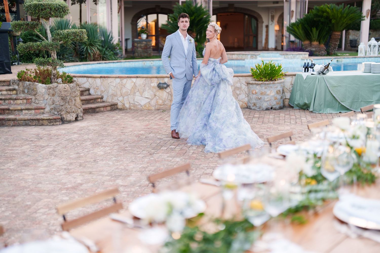Couple in blue wedding attire walking near fountain at elegant outdoor venue with decorated reception tables.