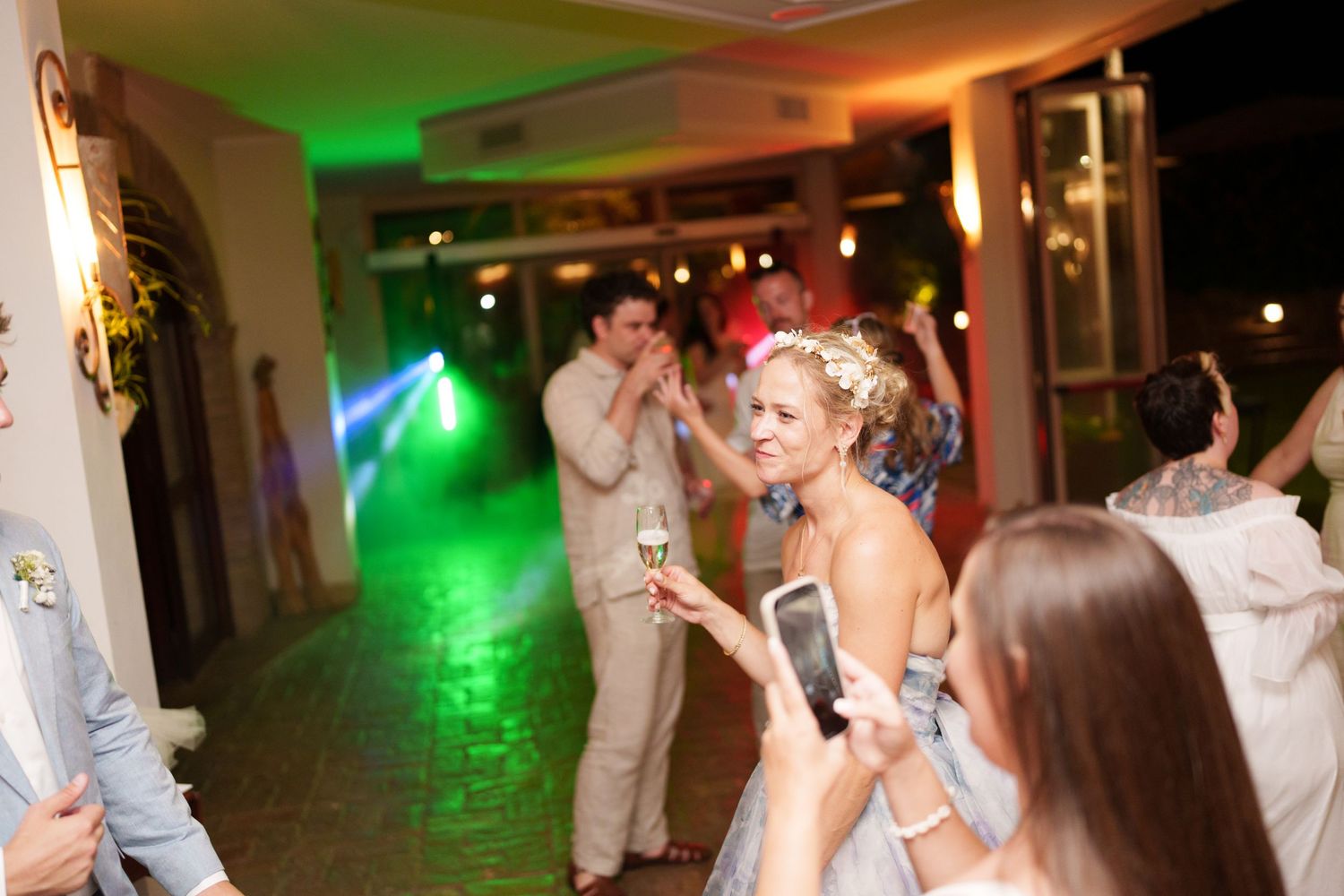 Guests dancing and celebrating at a wedding reception with colorful green and purple lights indoors.