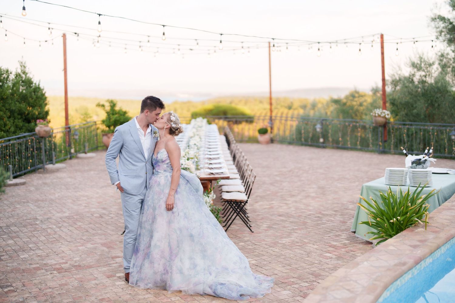 Bride and groom share a romantic moment at sunset outdoor wedding reception with long dining table and string lights.