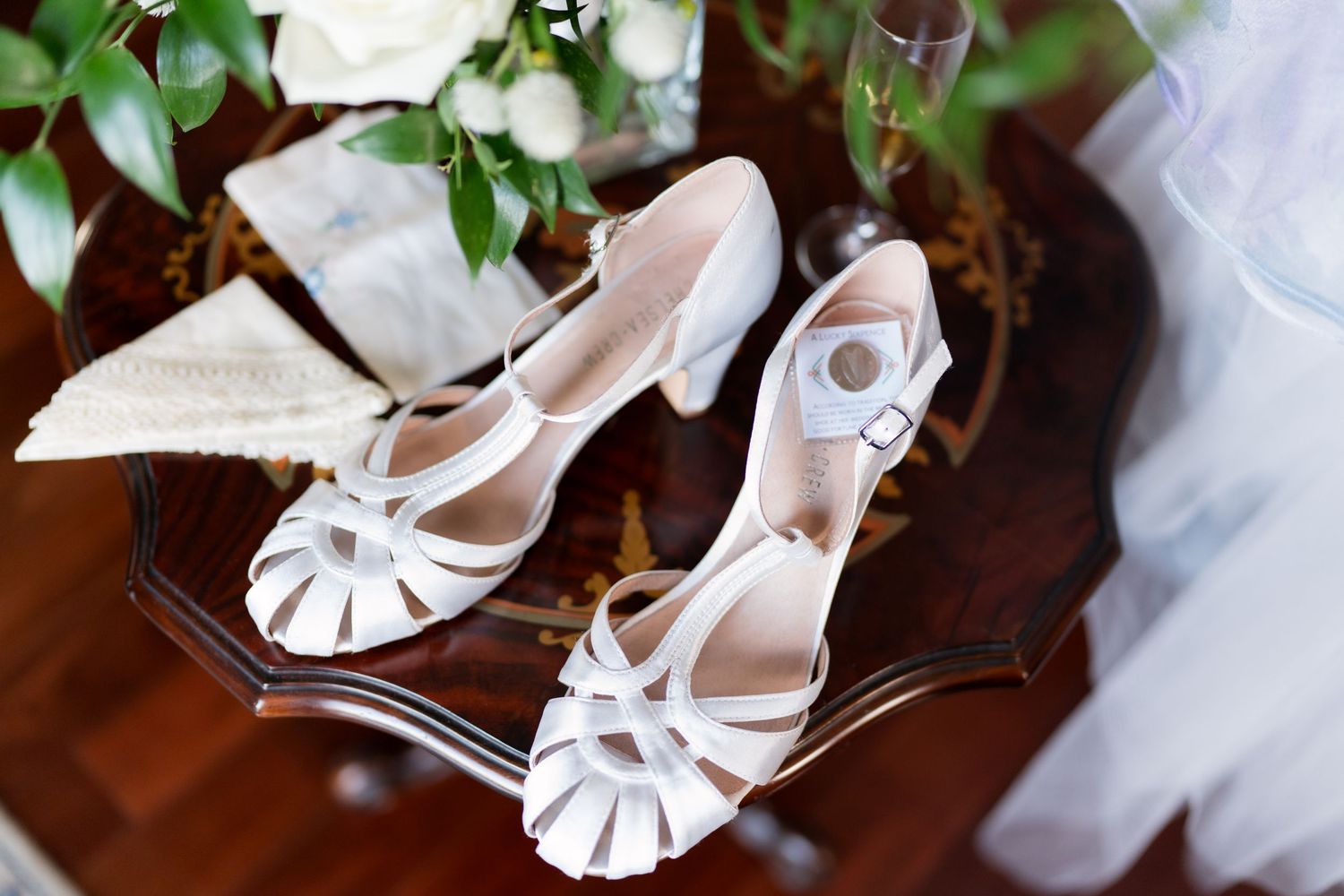 White strappy bridal heels with buckle detail displayed on a wooden table with greenery and wedding decor.