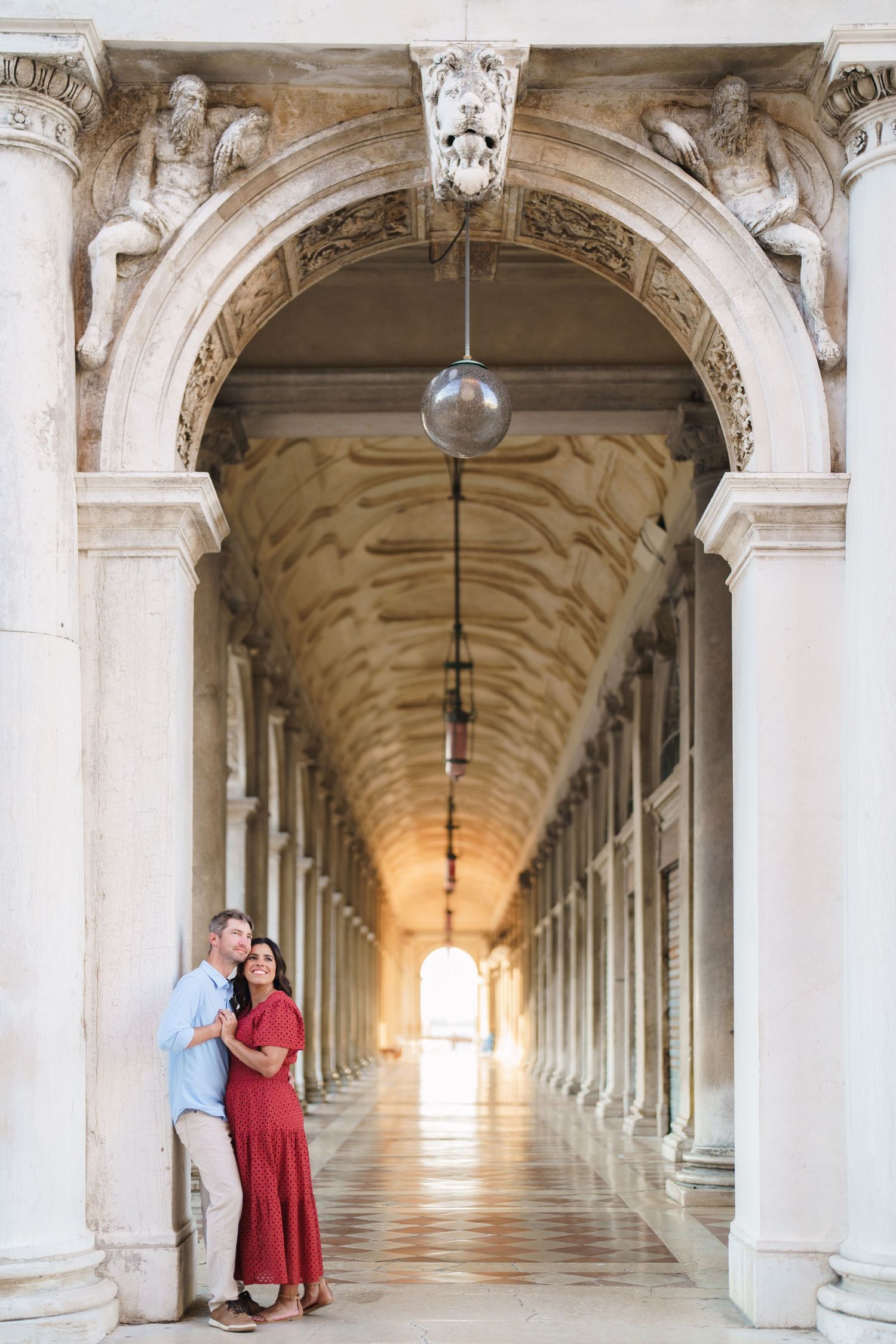 Couple embracing under ornate marble arches in a grand Venetian colonnade, woman in red dress, man in blue shirt.