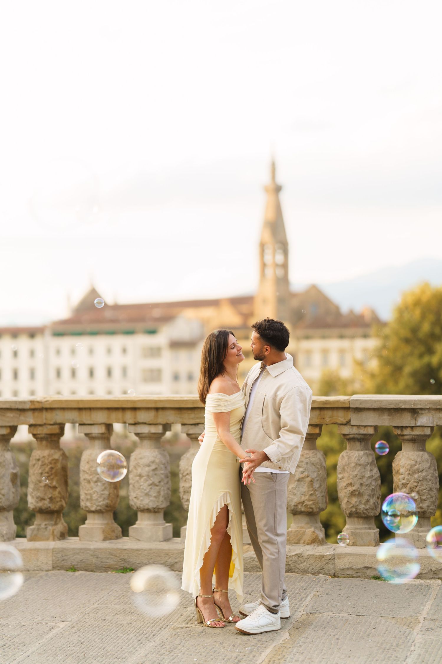 Romantic couple sharing a tender moment on a stone bridge with soap bubbles and European cityscape in the background.