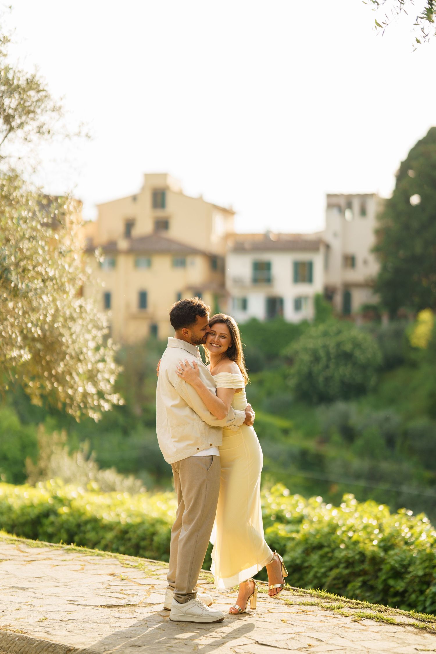 Romantic couple embracing in golden sunlight with lush greenery and Italian buildings in the background.