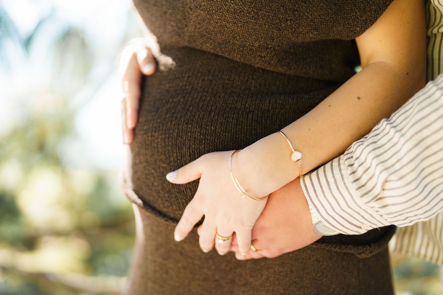 Close-up of couple holding hands over pregnant belly, wearing bracelets and rings outdoors.