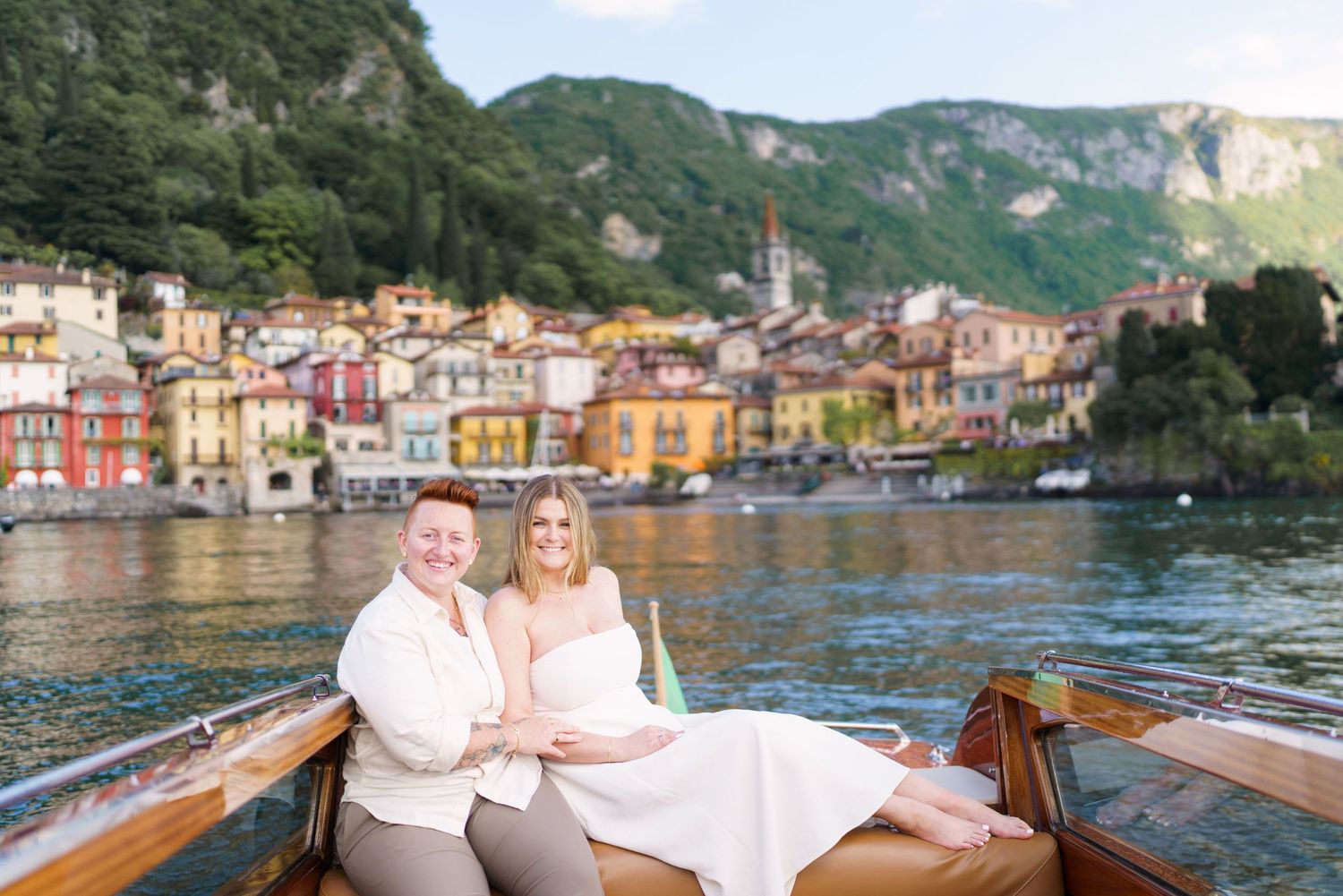 Couple smiling on a wooden boat with colorful Italian village and green hills on Lake Como in background.