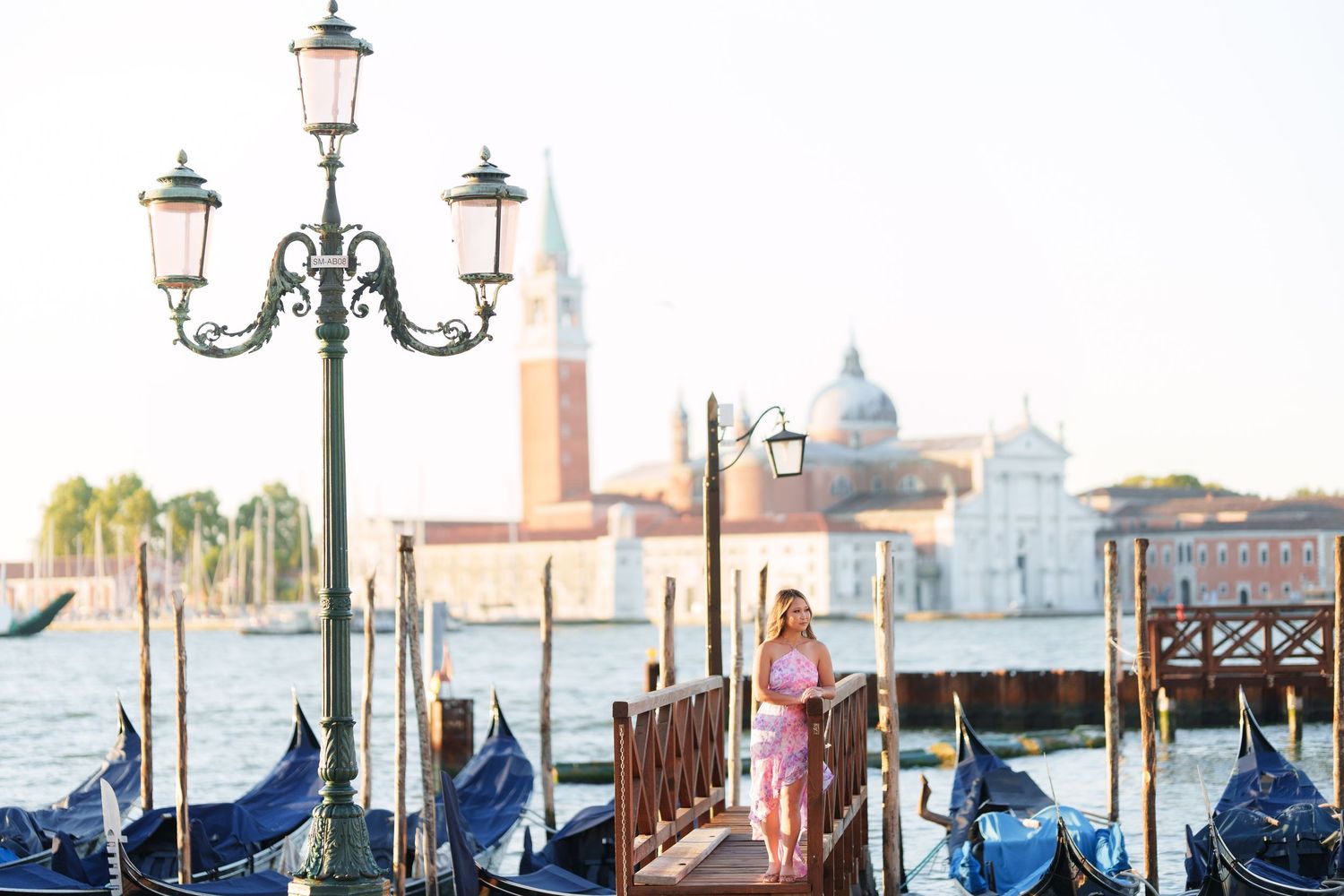Woman in pink dress walking on Venice gondola dock with San Giorgio Maggiore church and lagoon in background.