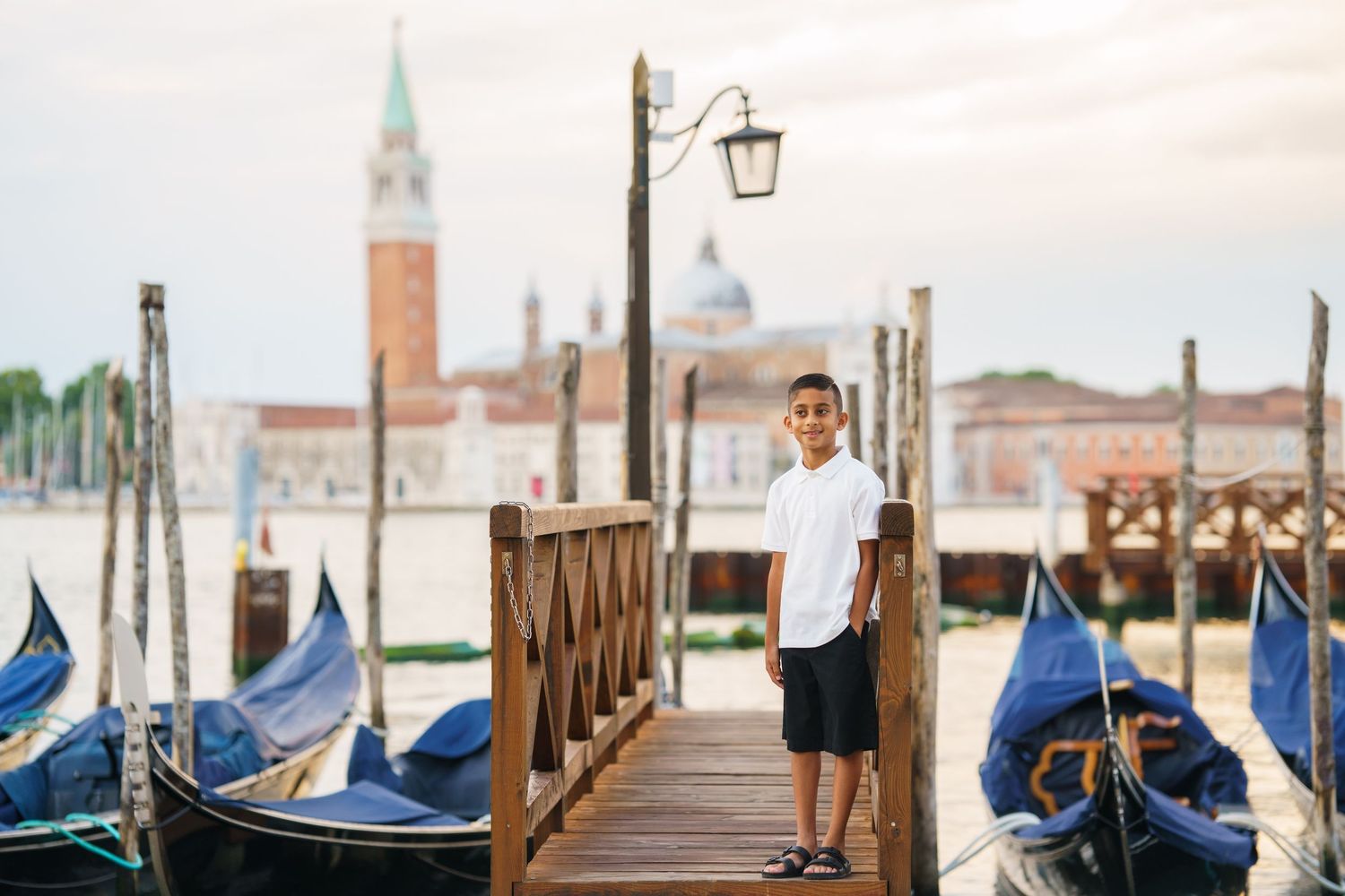 A boy stands on a wooden dock in Venice, Italy, surrounded by gondolas with San Giorgio Maggiore in the background.