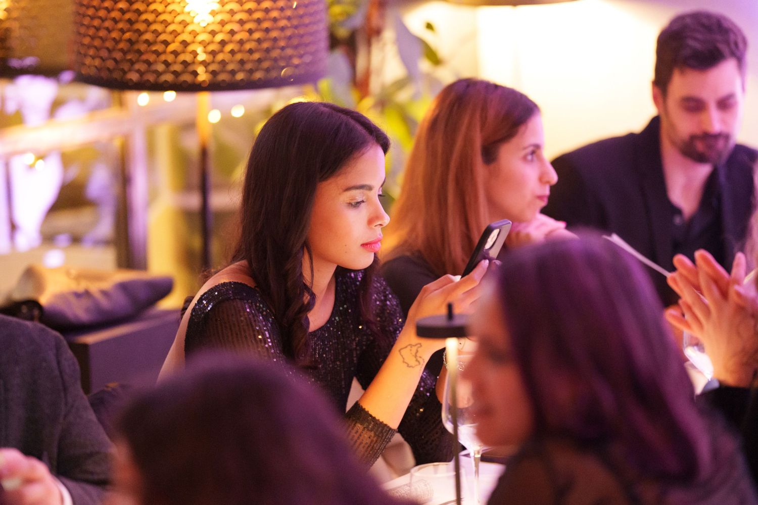 People socializing at a dimly lit restaurant, woman in black sequin dress using smartphone.