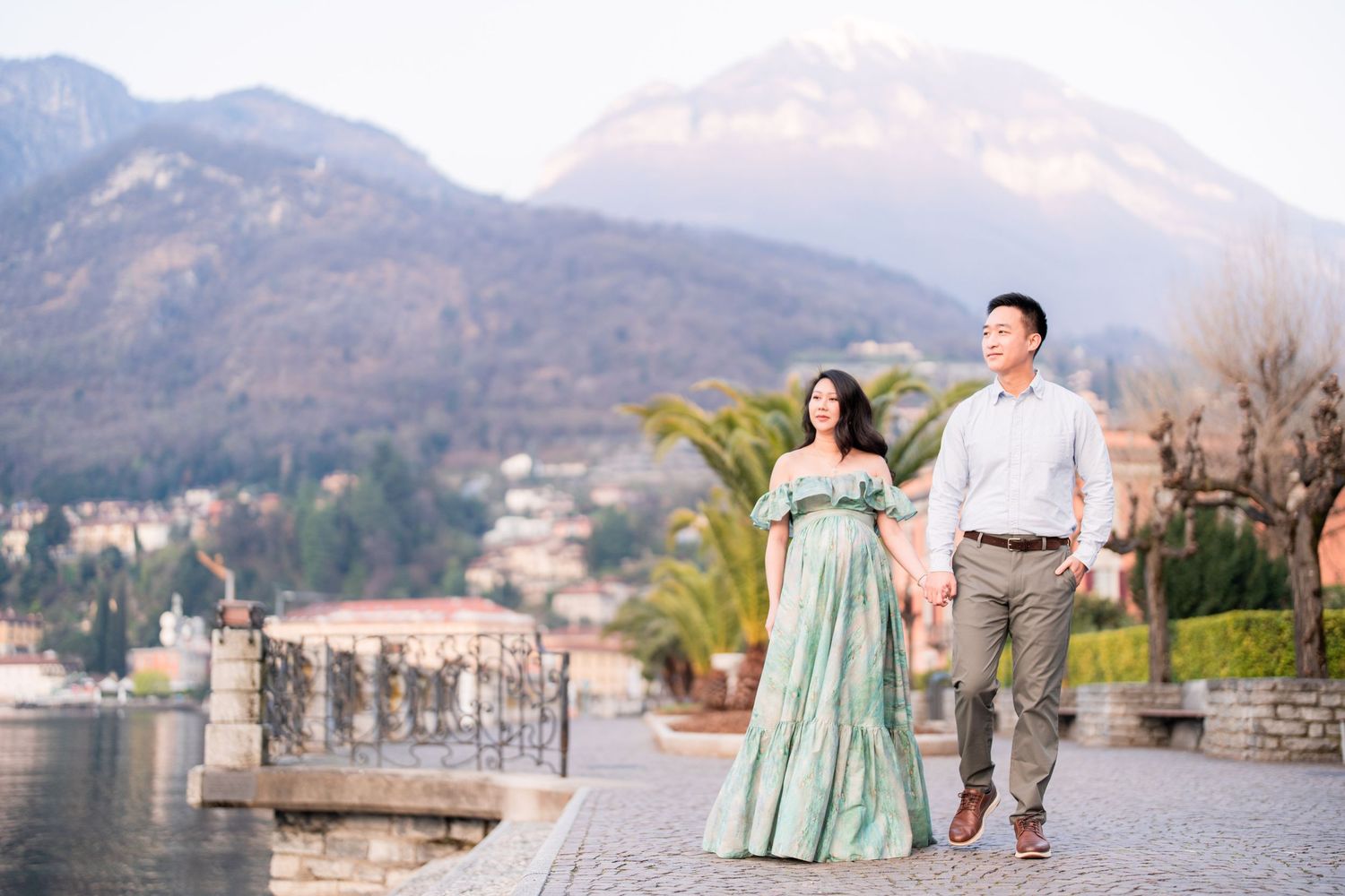 Couple holding hands walking along lakeside promenade with mountain backdrop at Lake Como, Italy.