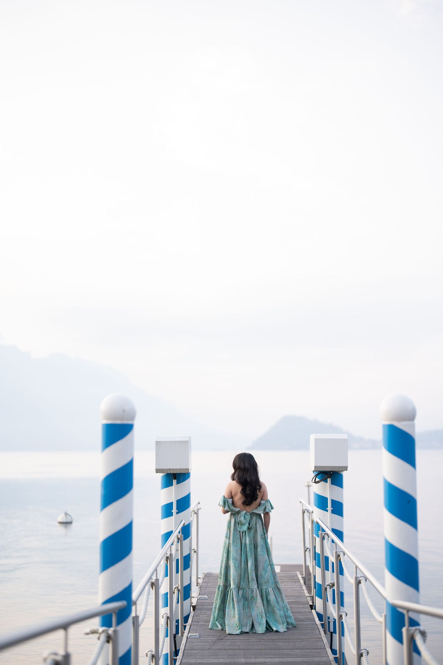 Woman in teal off-shoulder gown standing on pier with blue striped posts overlooking calm lake and mountains.