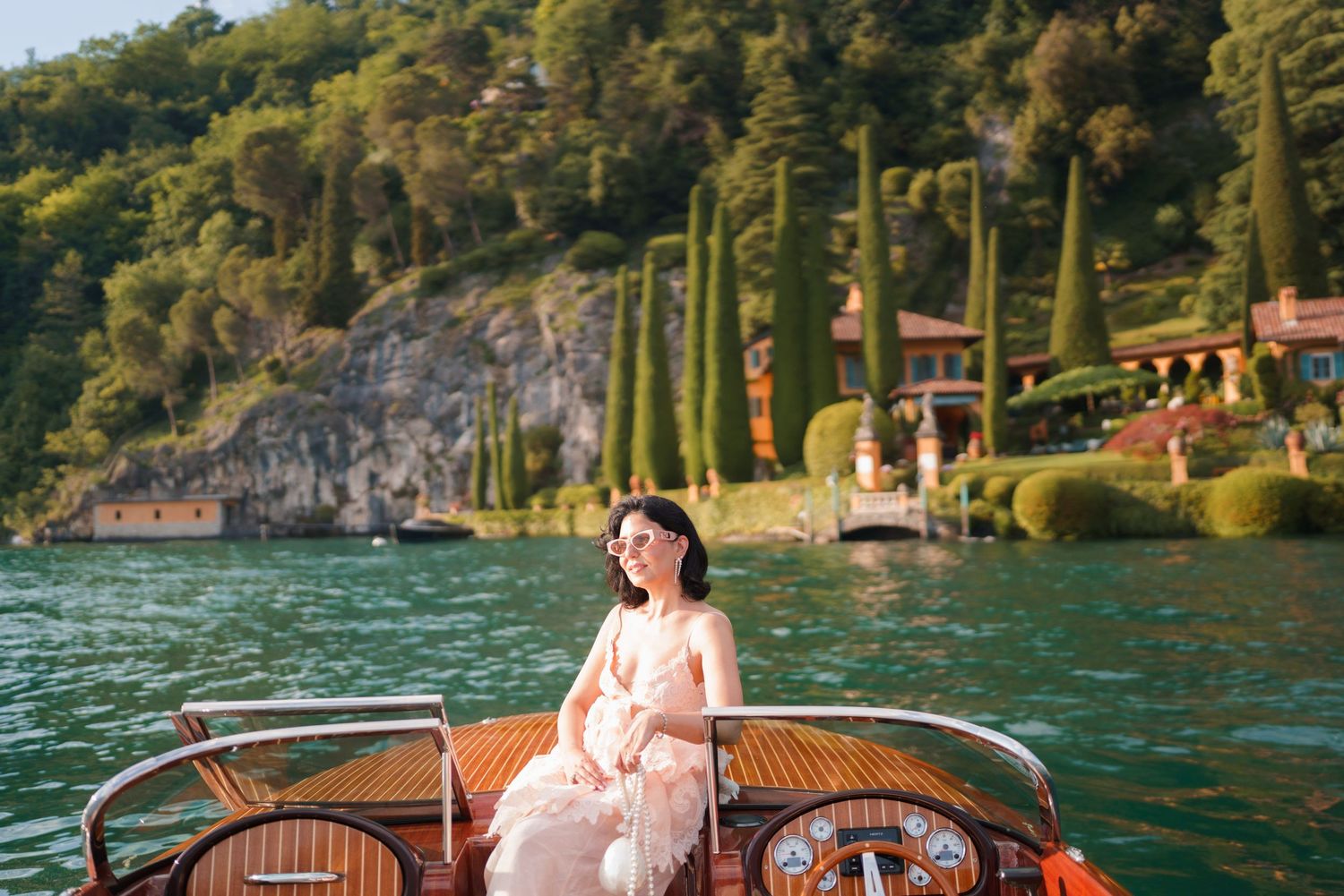Woman in pink dress sitting on a wooden boat on Lake Como with lush greenery and villa in background.