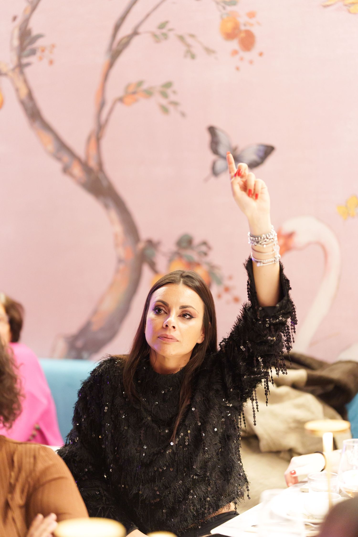 Woman in black sequin top raising finger at elegant restaurant with pink floral mural backdrop.