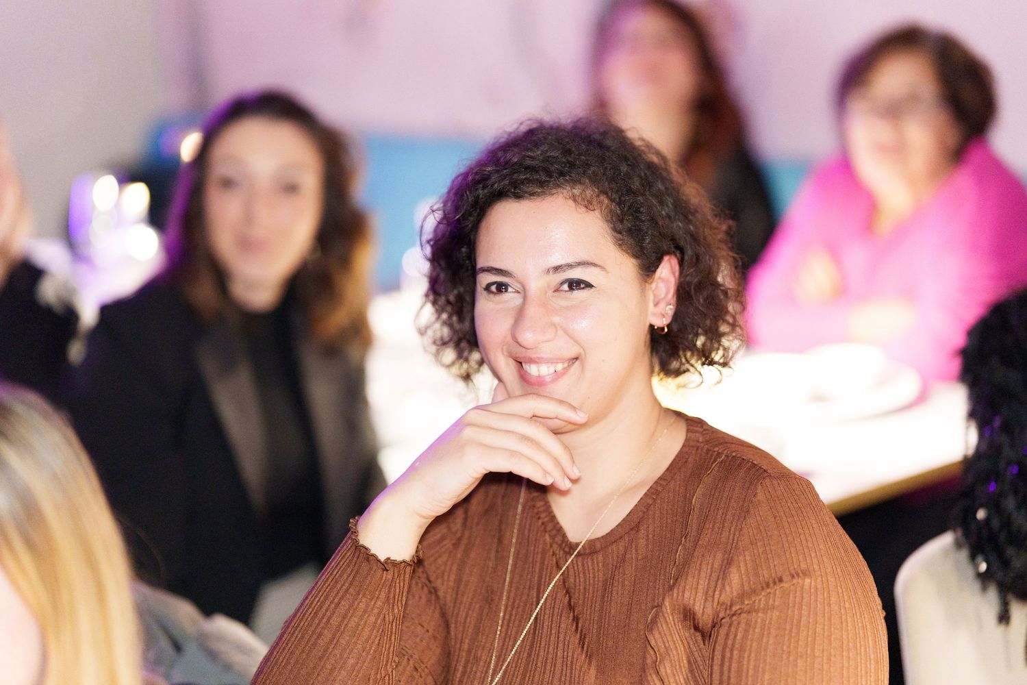 A young woman with curly hair smiles warmly at an indoor gathering, resting her chin on her hand.