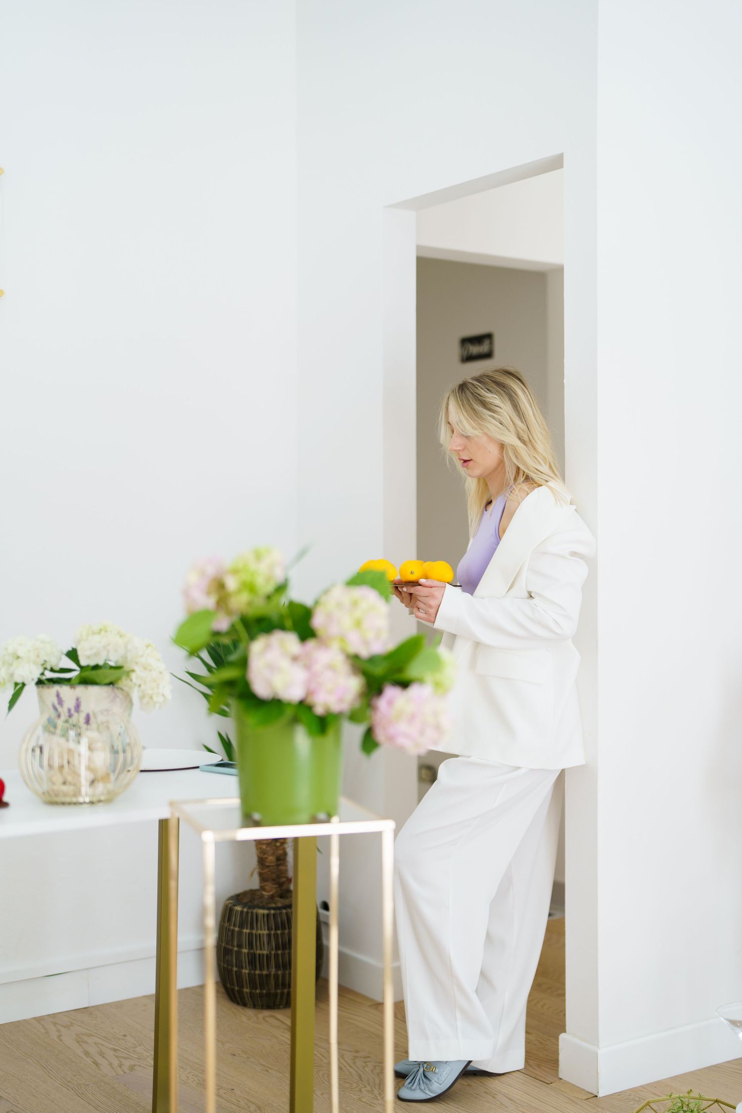 Blonde woman in white suit holding yellow cup near floral arrangements in bright modern interior.
