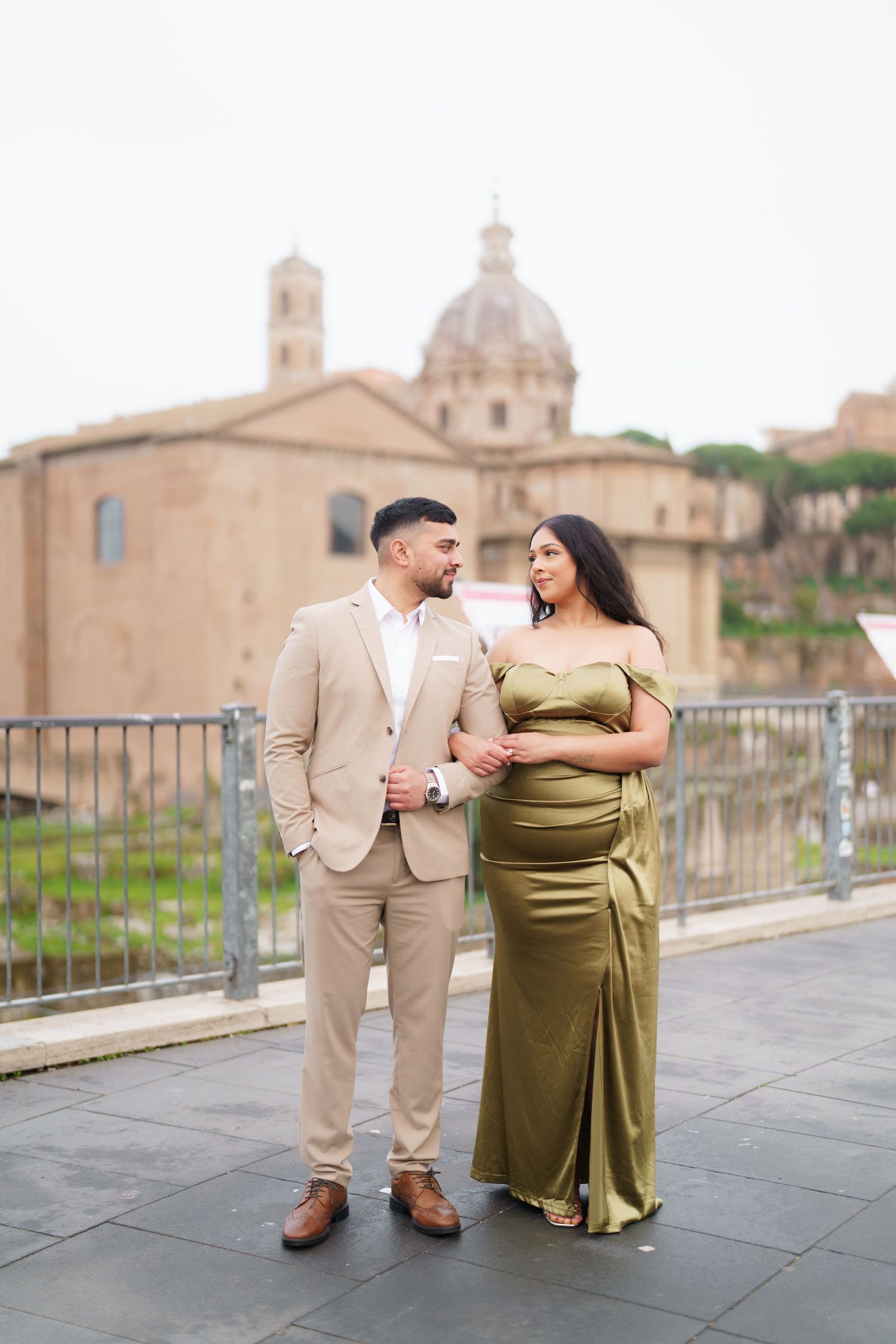 Elegant couple posing in Rome, woman in olive satin gown, man in beige suit, with historic Roman architecture behind them.