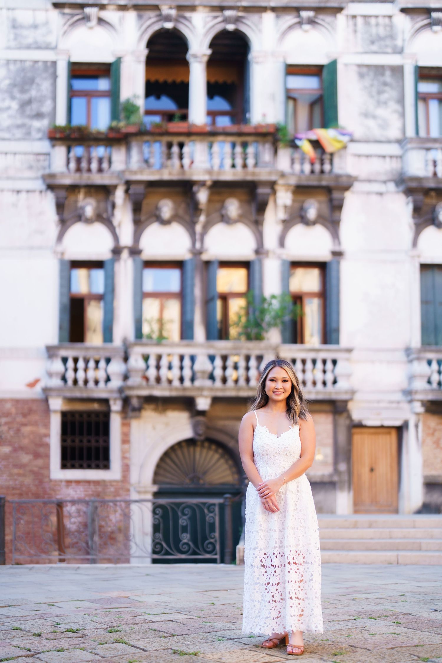Young woman in white lace dress poses before ornate Venetian building with balconies in Venice, Italy.
