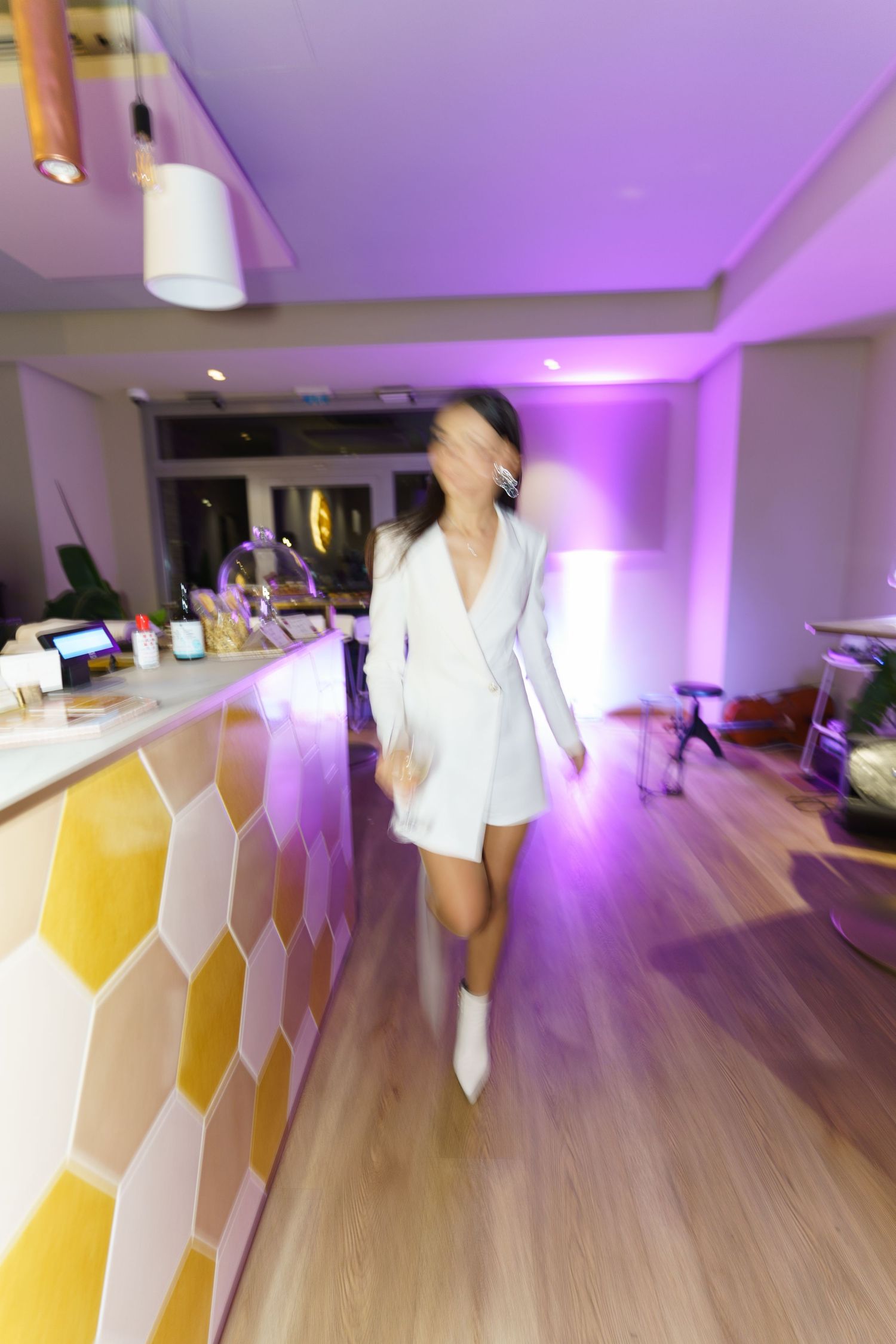 Woman in white dress walking through a modern bar with purple lighting and colorful honeycomb tile accents.