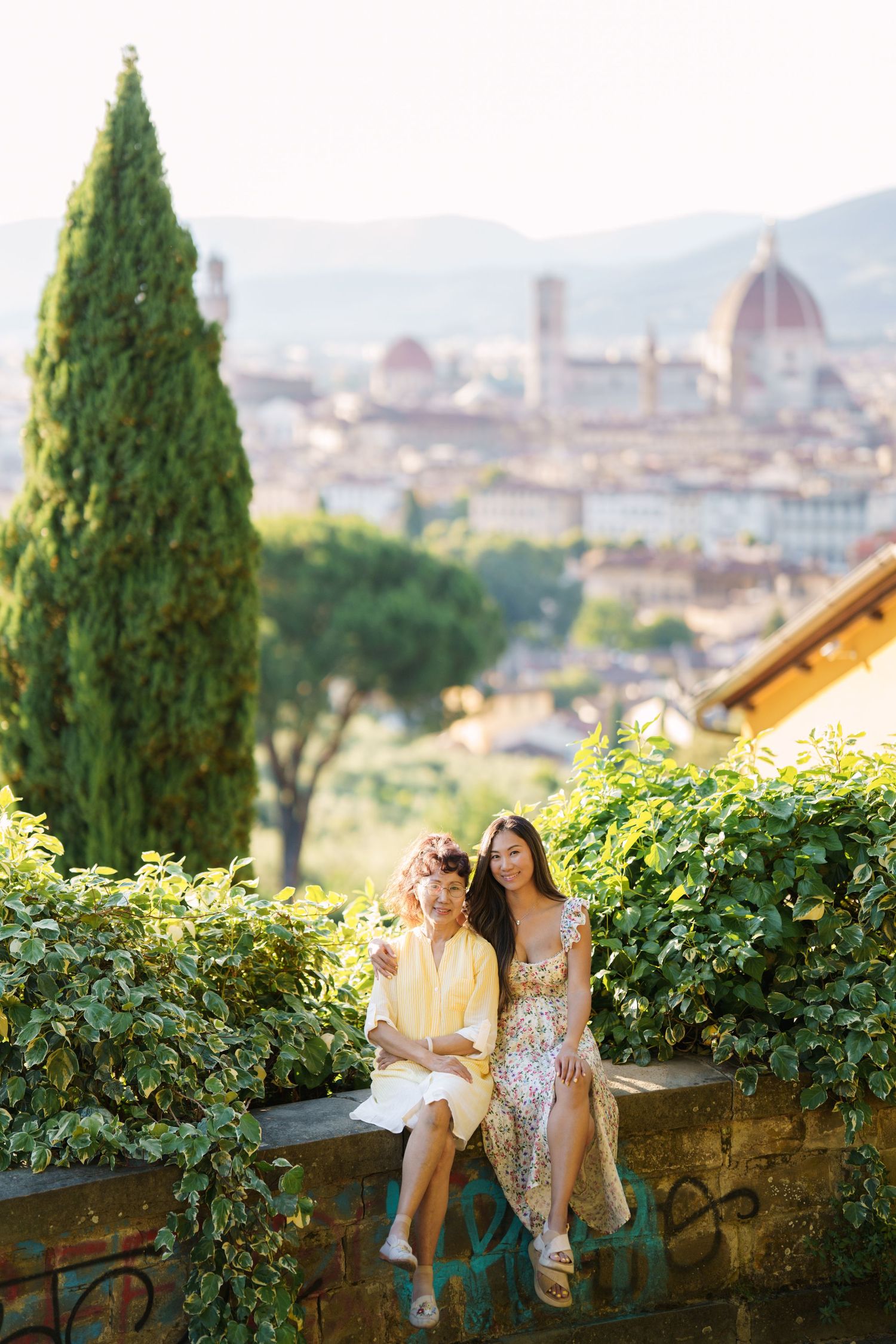 Two women in summer dresses sit on a stone wall with Florence family vacation photographer italy
