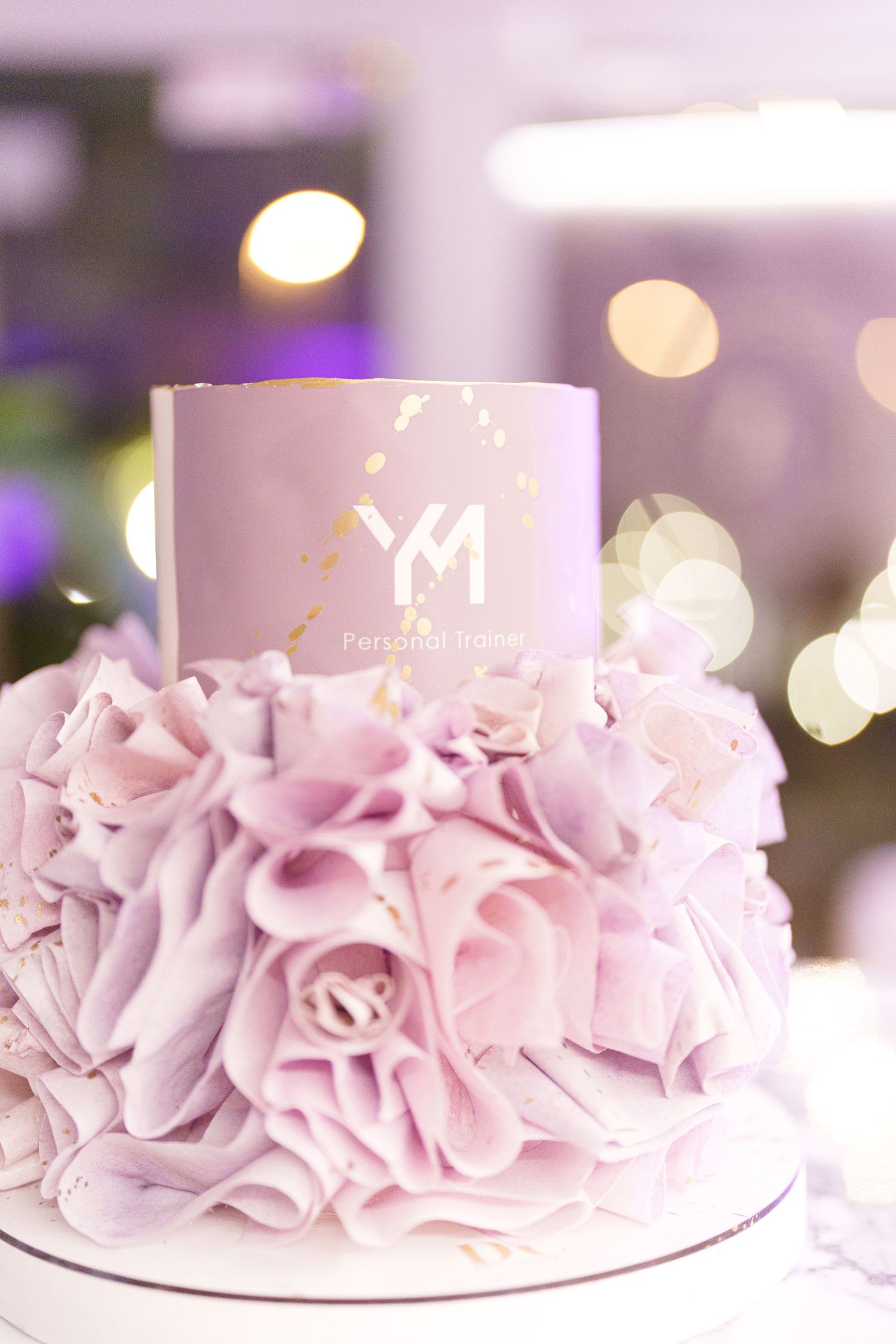 Elegant two-tier lavender ruffle cake with YM Personal Brand gold logo topper on a white base.