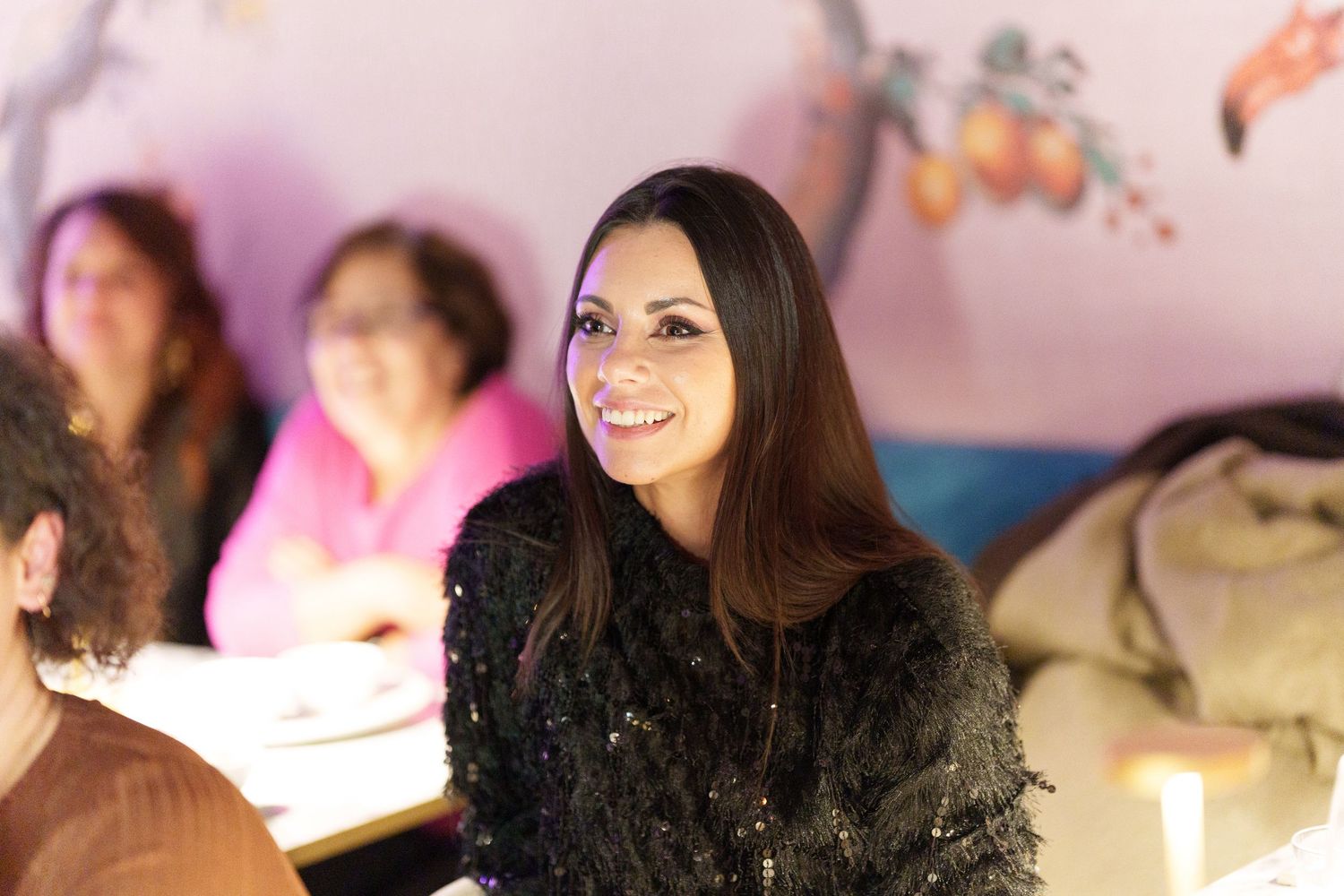 Smiling young woman with long dark hair wearing a black sequin jacket seated at a table indoors.
