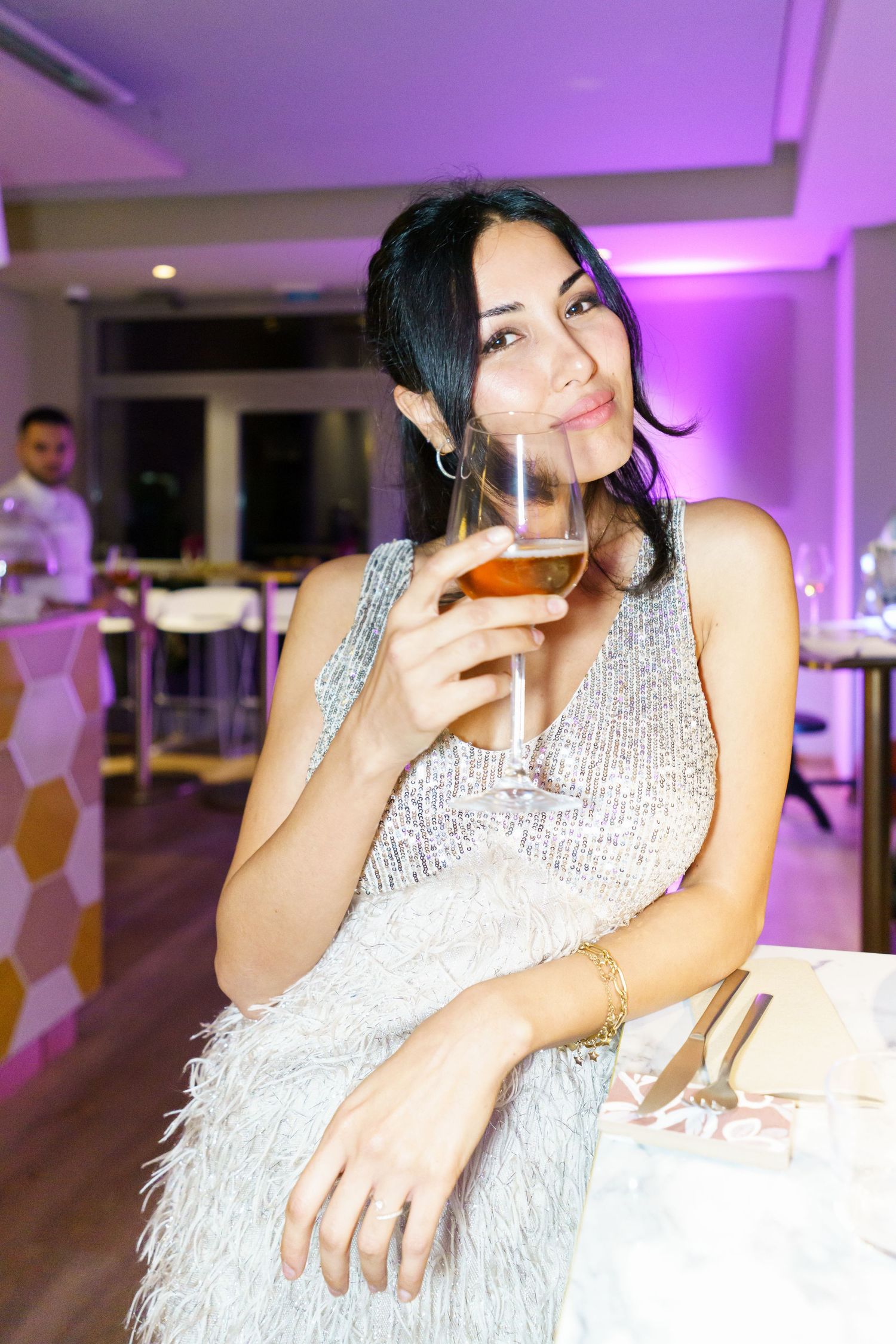 Elegant woman in silver fringe dress holding a champagne glass at a upscale restaurant with purple ambient lighting.
