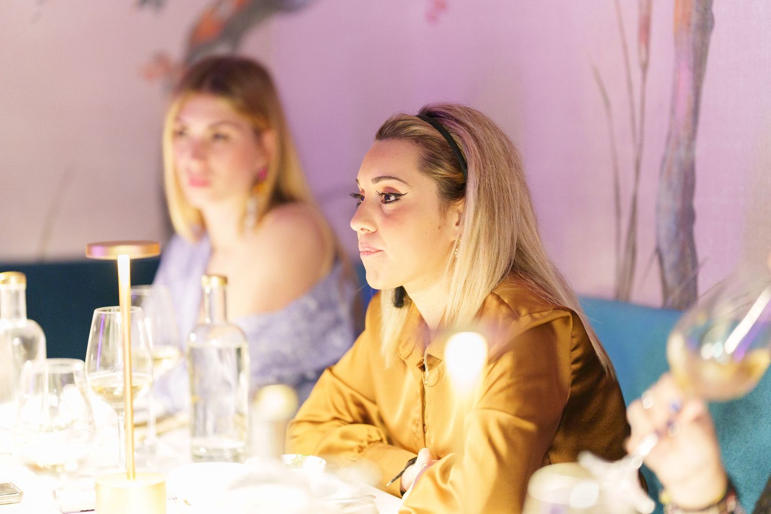 Two women sitting at a dining table with glassware, one in a gold jacket attentively listening at an elegant restaurant.